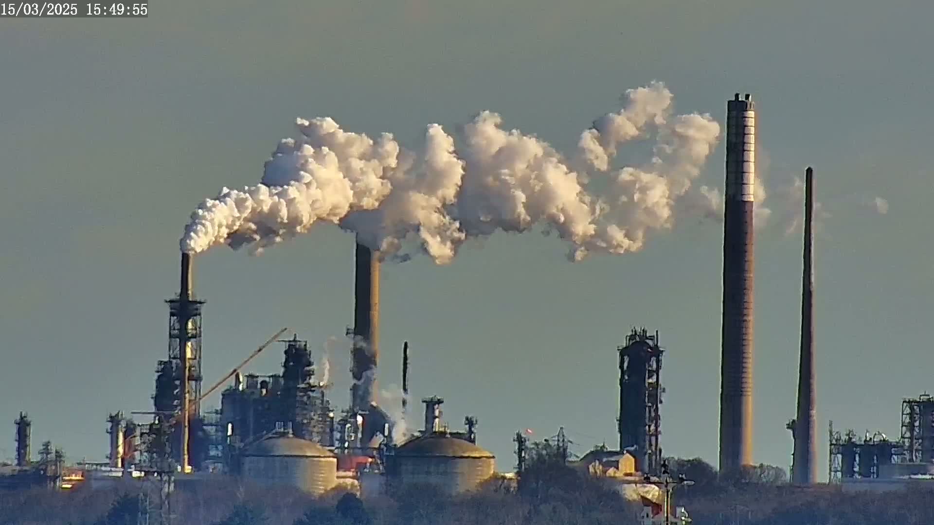 White smoke billows from several tall industrial smokestacks of a refinery under a clear, light-grey sky.