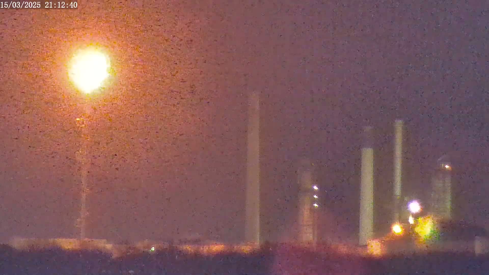 A bright light shines over an industrial area at night, with several tall structures visible in the hazy, dusky atmosphere.