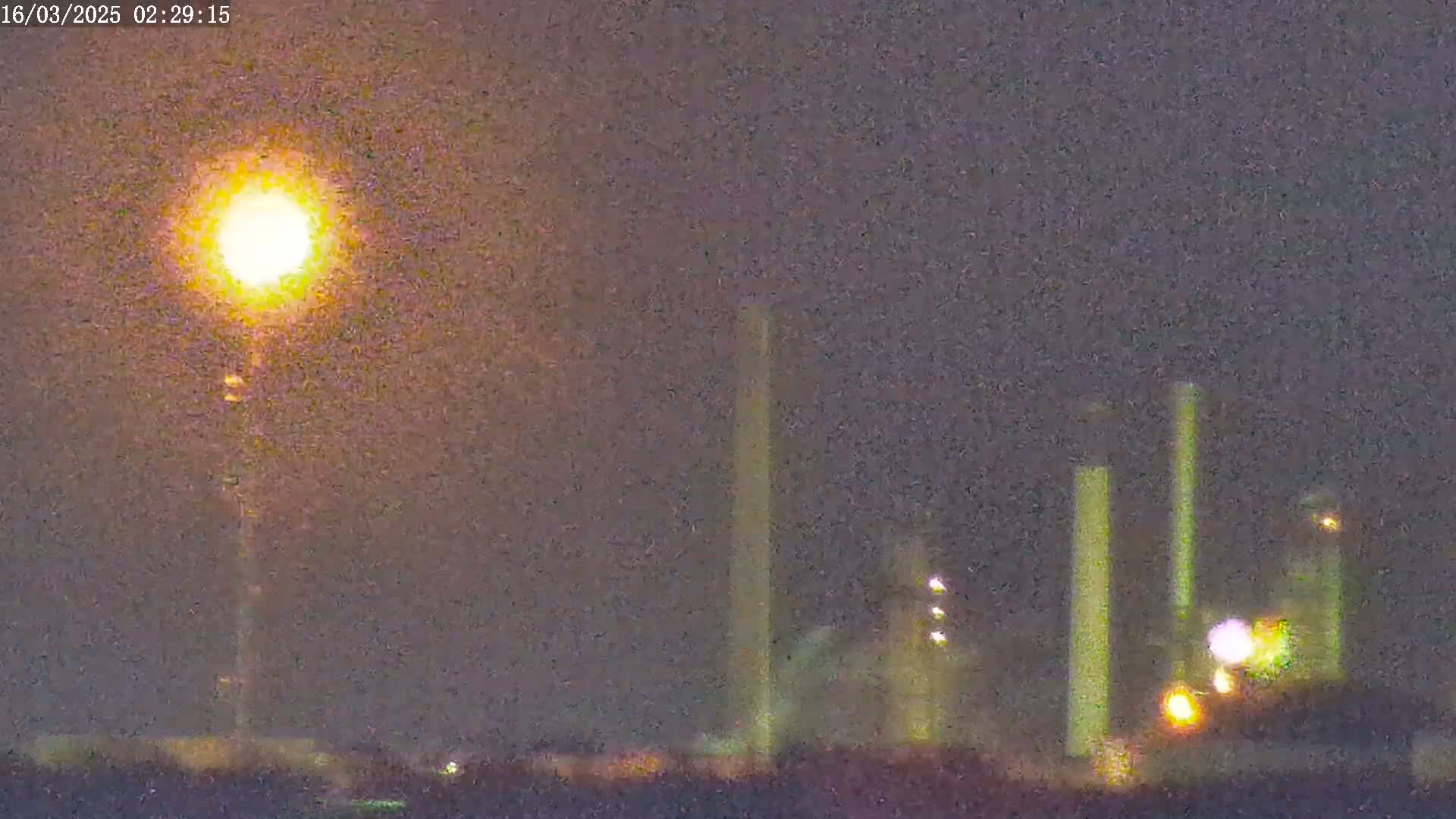 A bright light illuminates a nighttime scene featuring tall industrial structures, possibly smokestacks, under a hazy sky.