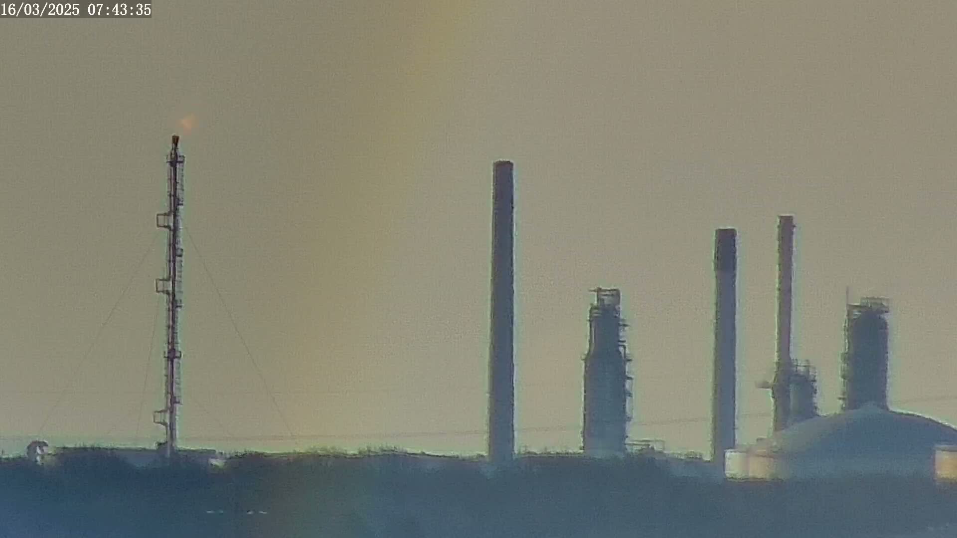 A refinery with several tall smokestacks and a flaring gas tower is visible against a hazy, light-brown sky.