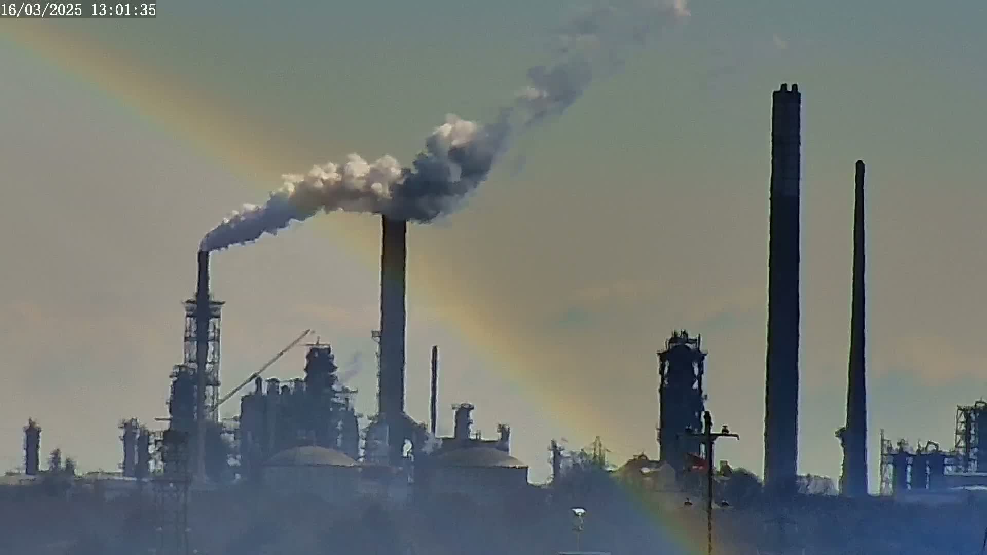 A large industrial complex with several tall smokestacks emitting smoke is visible against a hazy sky, partially obscured by a faint rainbow.