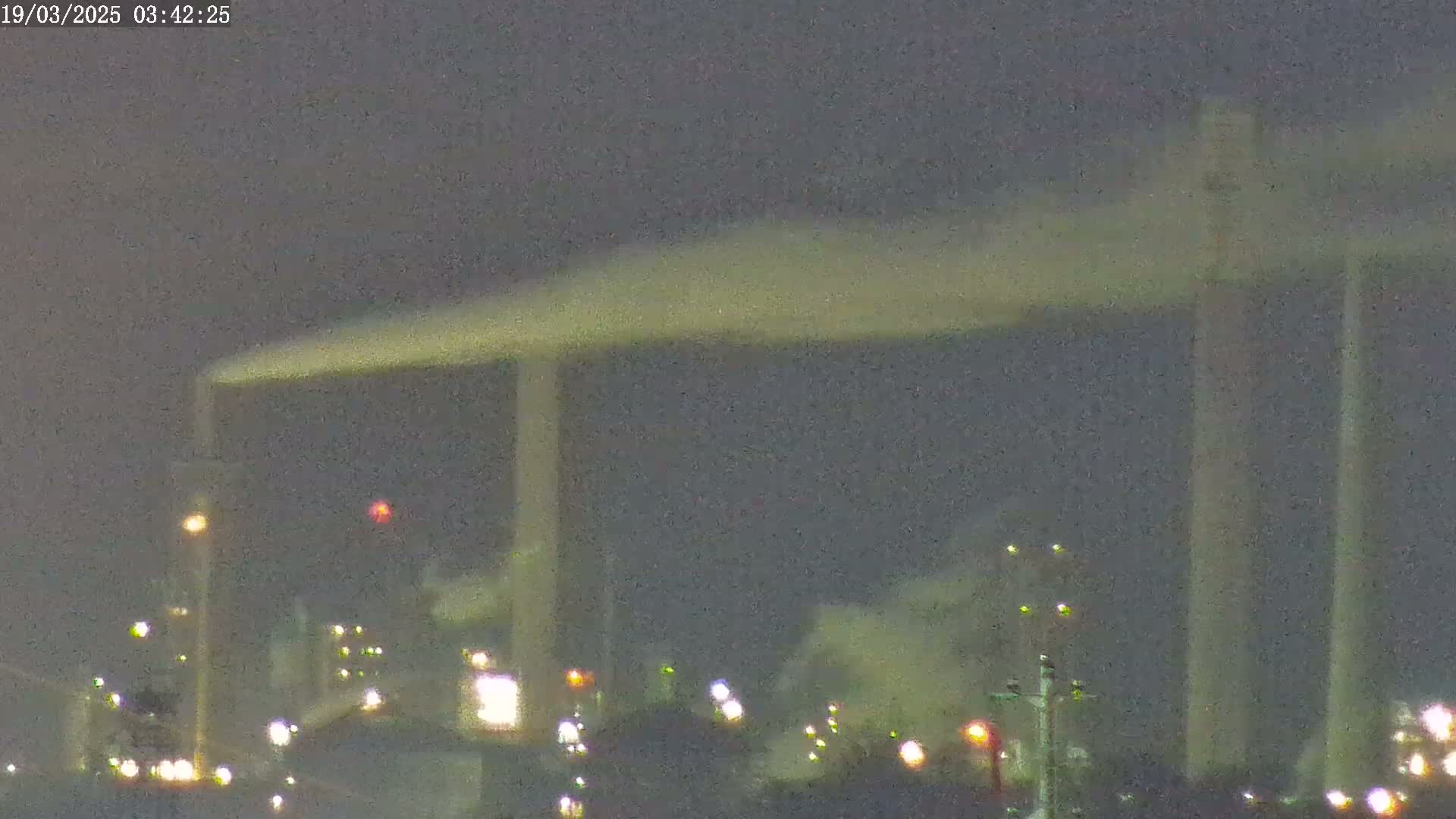 At night, a large industrial complex with tall smokestacks emitting plumes of smoke is illuminated by scattered lights under a dark sky.