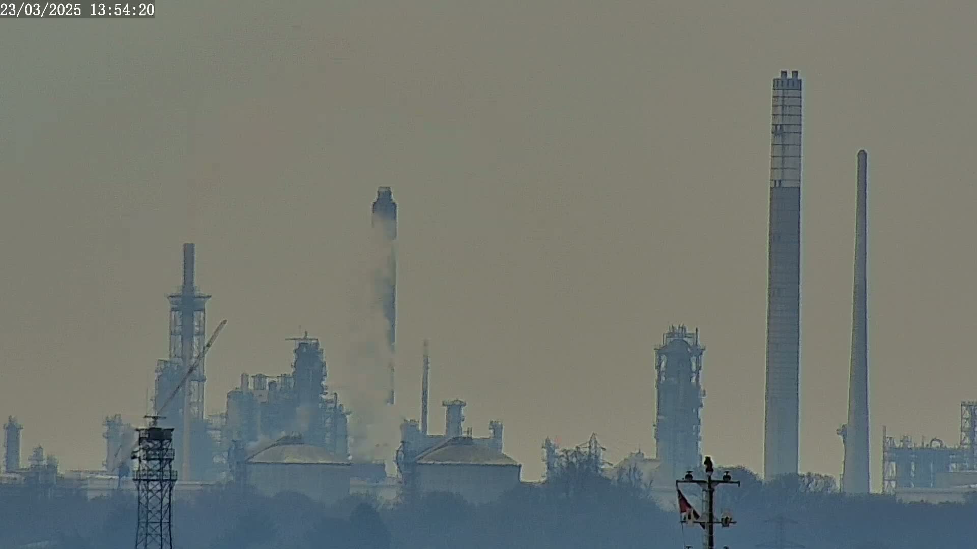 A hazy view of an industrial complex with tall smokestacks emitting plumes of smoke, situated behind a line of trees.