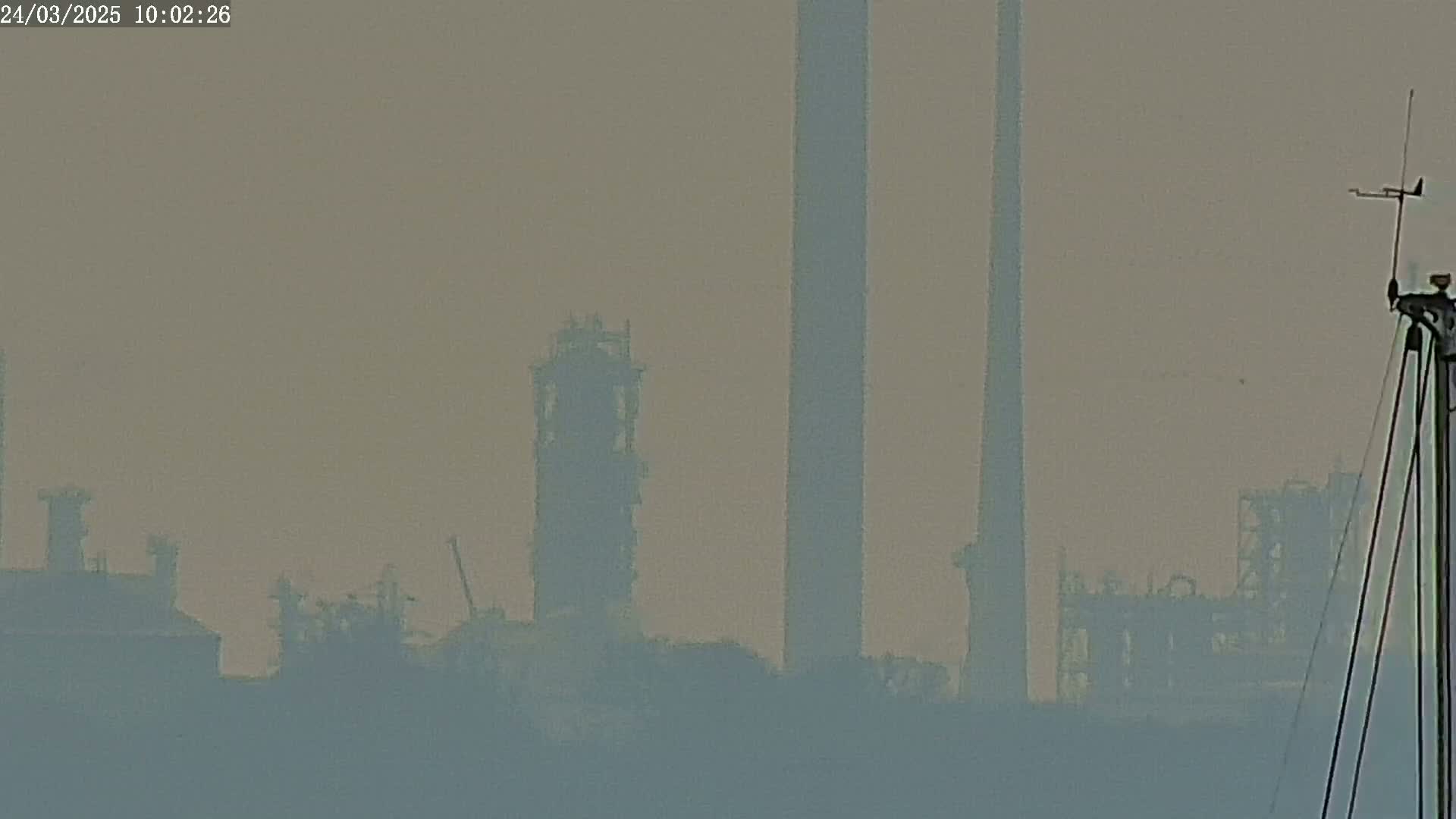 A hazy, distant view of industrial structures, including tall smokestacks and refinery buildings, is visible from a vantage point that includes a portion of a weather vane.