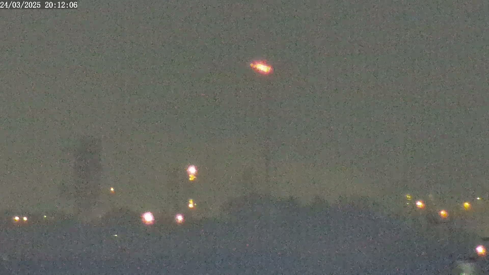 A blurry, nighttime image shows a bright orange light in the sky above a distant line of faint lights on the ground under a dark, overcast sky.