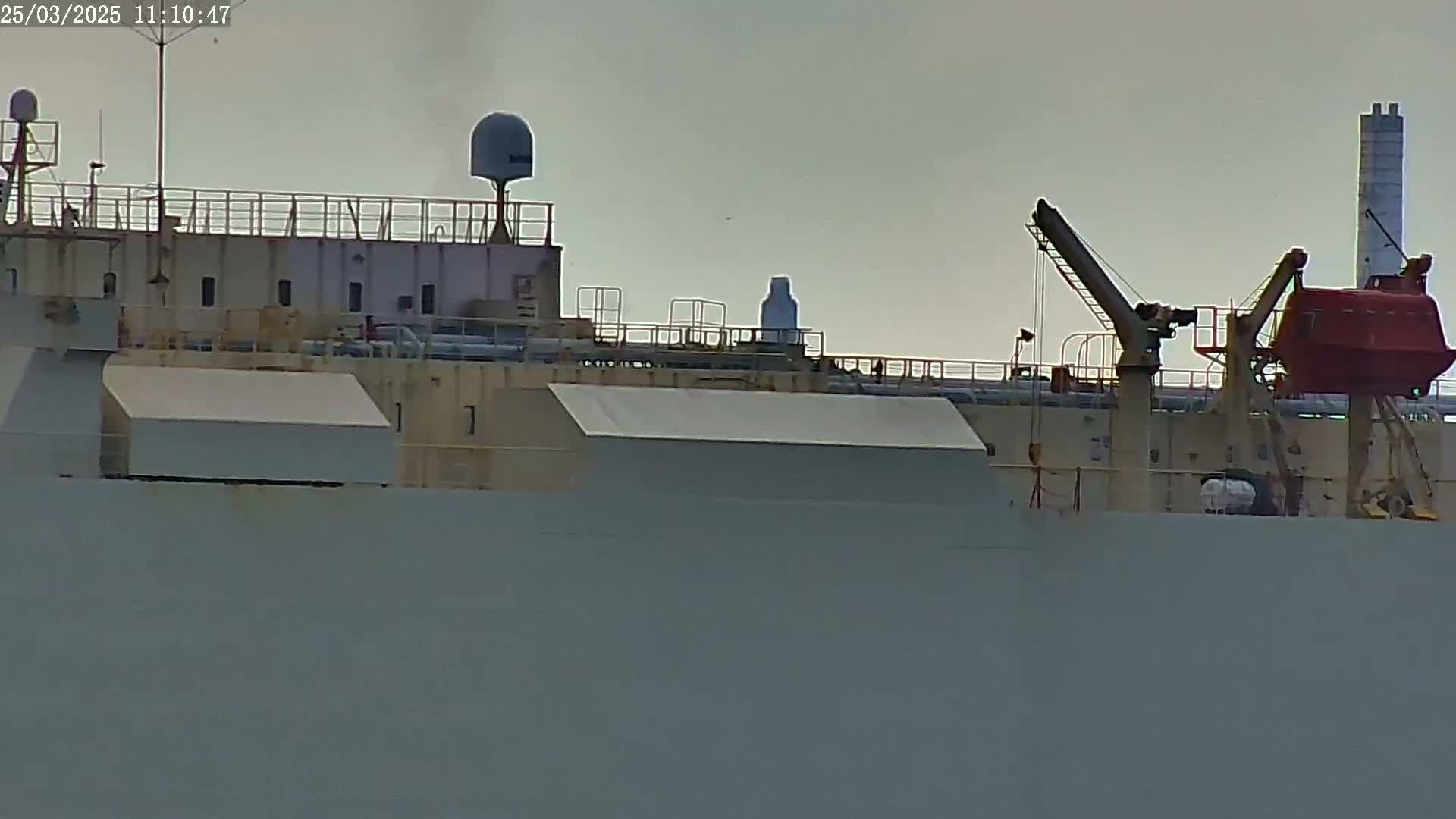 The upper deck of a large ship is shown under overcast skies, featuring various equipment including satellite dishes, railings, cranes, and a lifeboat.