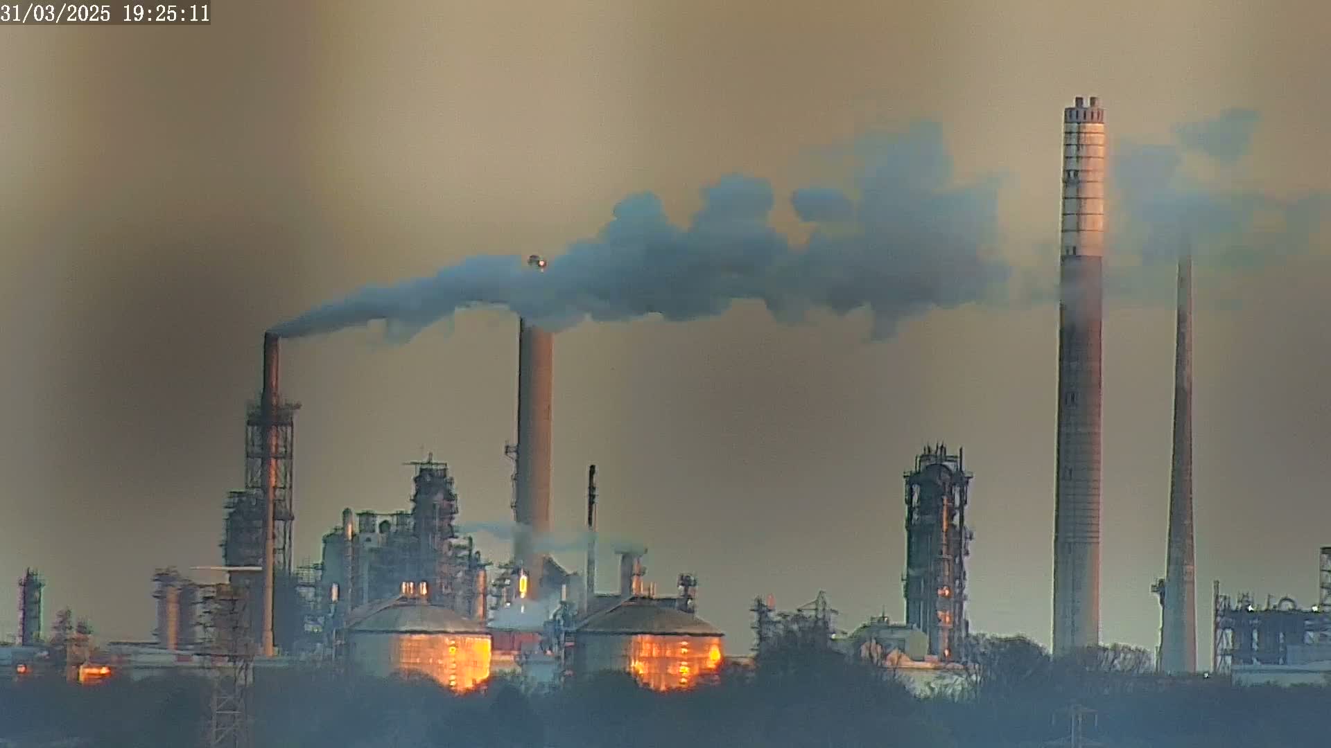 A large industrial complex with numerous smokestacks emitting plumes of smoke into a hazy, dusky sky.