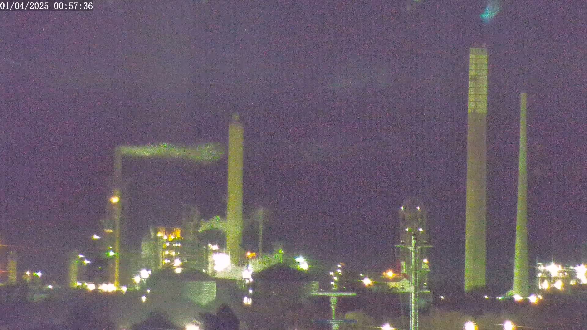 At night, under a dark sky, an industrial complex with tall smokestacks emits a plume of smoke.