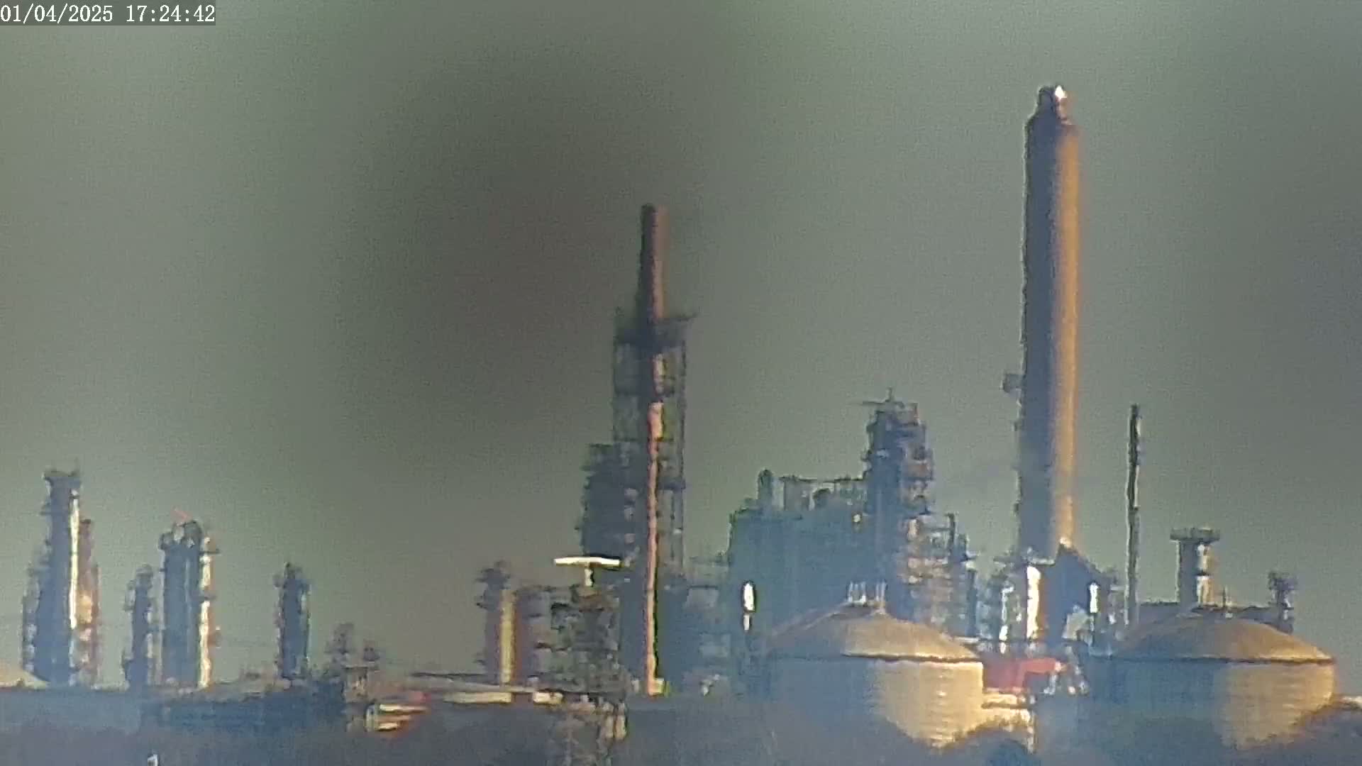 A hazy view shows an industrial refinery with several tall smokestacks and large structures under a muted sky.