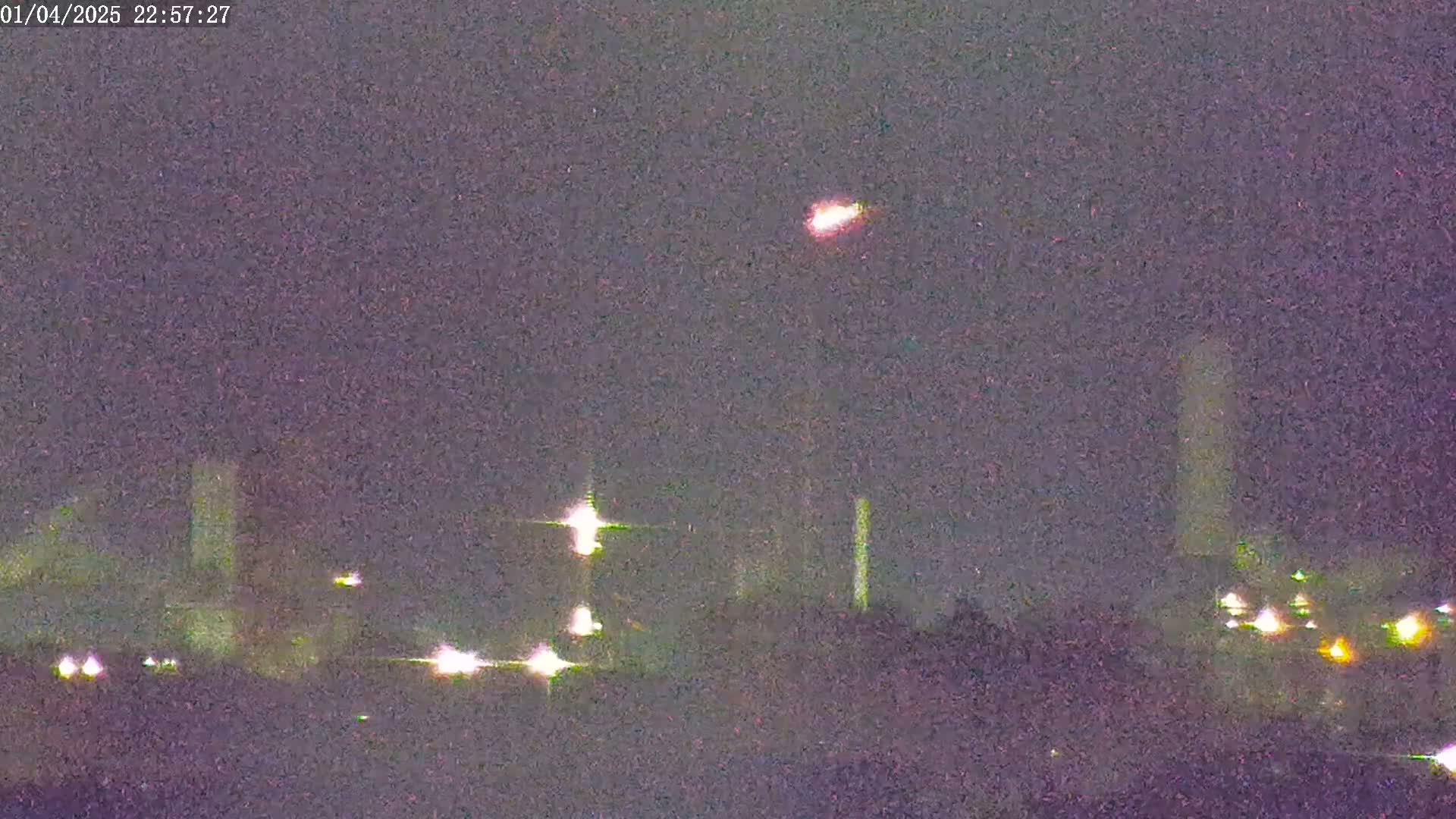 A nighttime long shot shows an industrial area with lights and tall structures under a dark, possibly cloudy sky, with a bright light streaking across the sky.