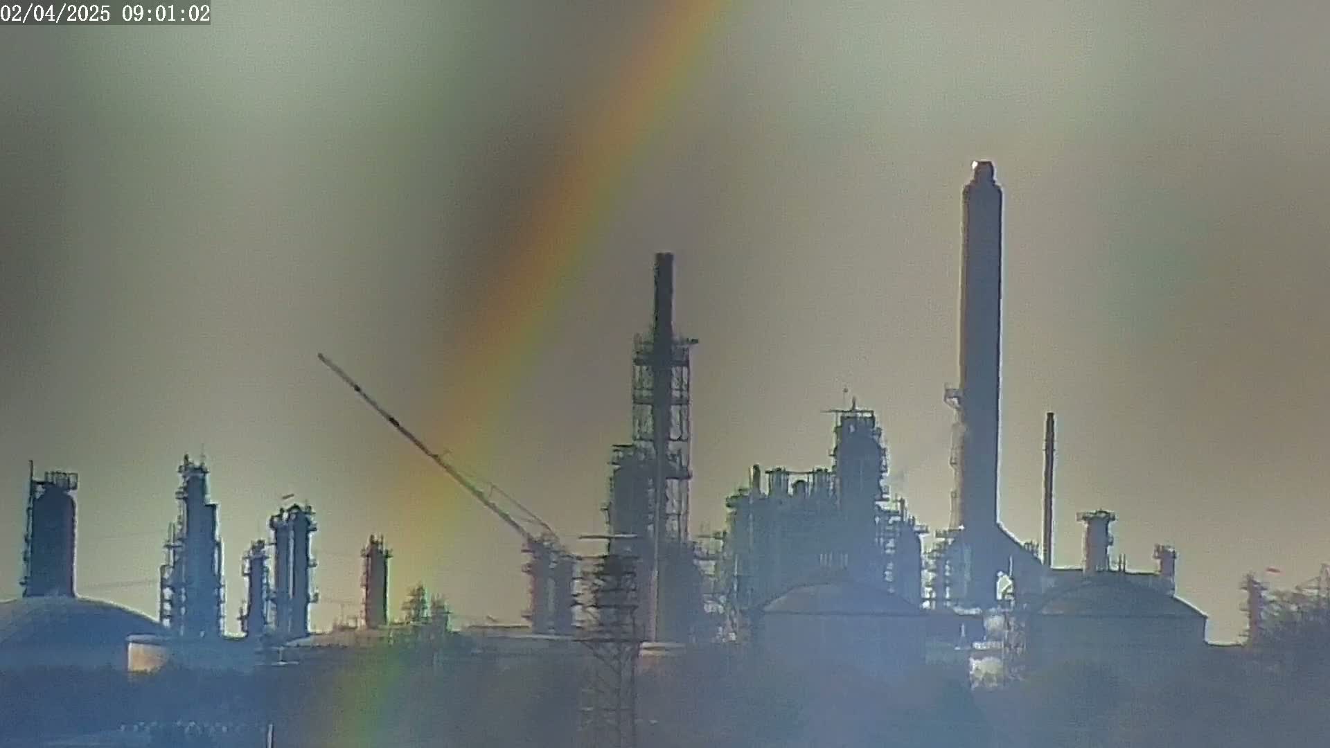 A large industrial plant is visible on a hazy day, with a partial rainbow in the sky.