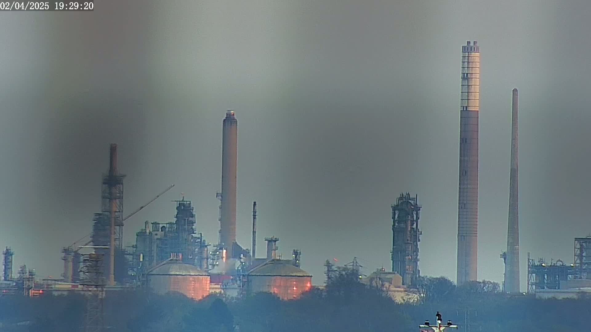 A hazy view of an industrial refinery complex with tall smokestacks, partially obscured by trees in the foreground.