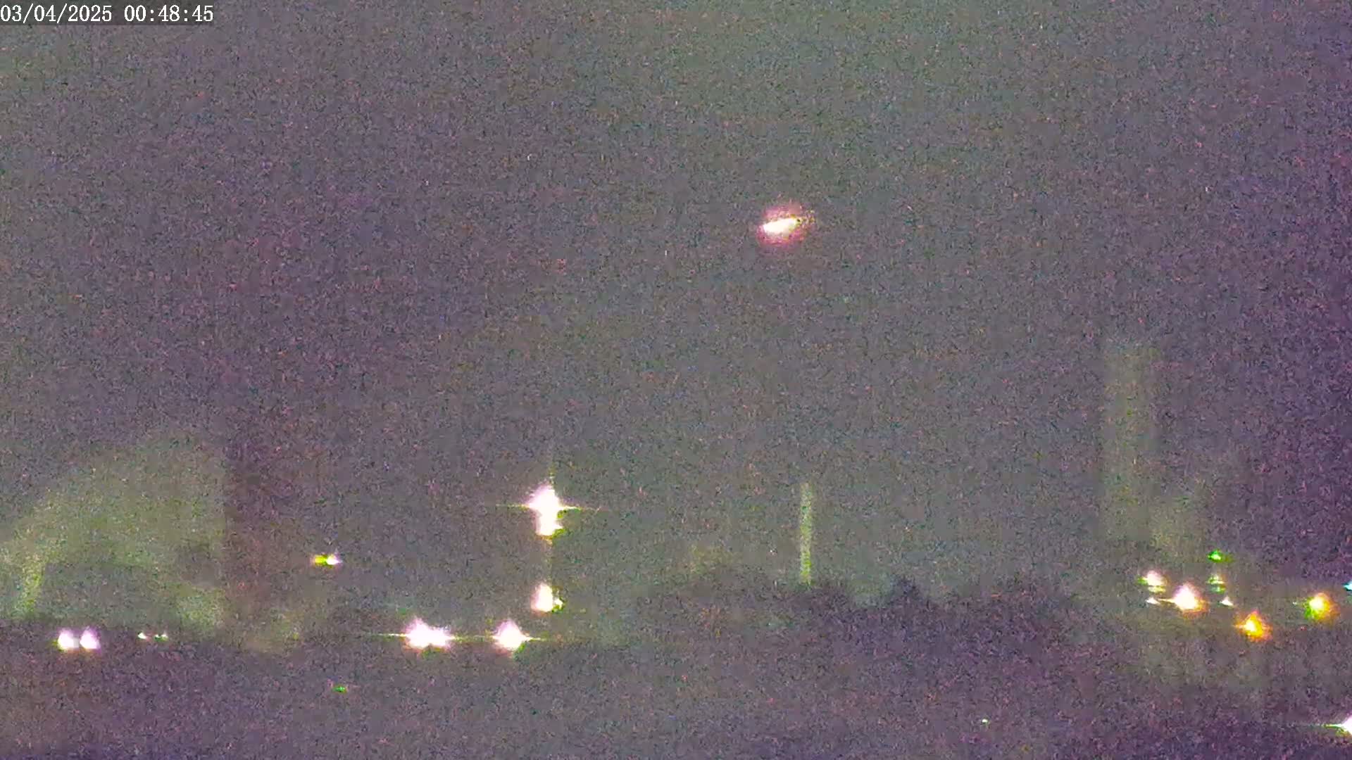 A nighttime long shot shows an industrial area with lights illuminating the scene under a dark sky with a single, distant light source.