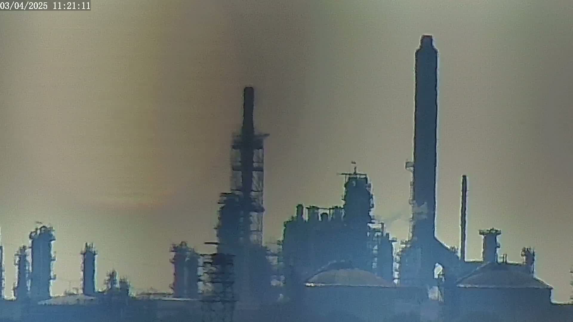A hazy view of an industrial refinery complex with tall smokestacks under a muted, light-brown sky.