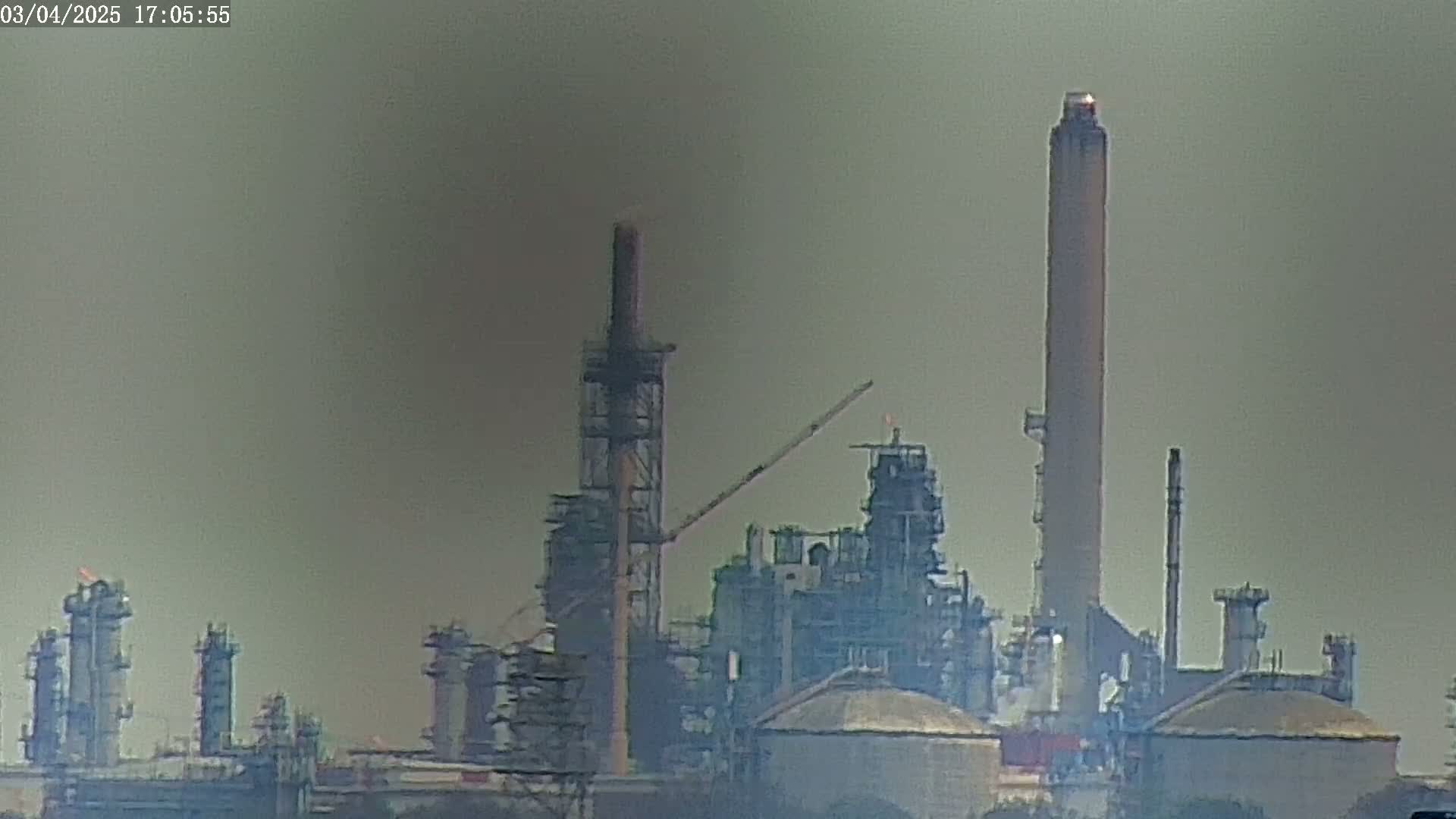 A hazy view of an industrial refinery with numerous tall structures and smokestacks under an overcast sky.
