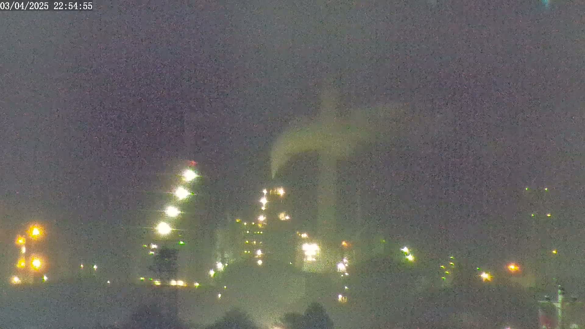 A nighttime view of an industrial complex with multiple illuminated buildings and a plume of smoke or steam under a dark, hazy sky.