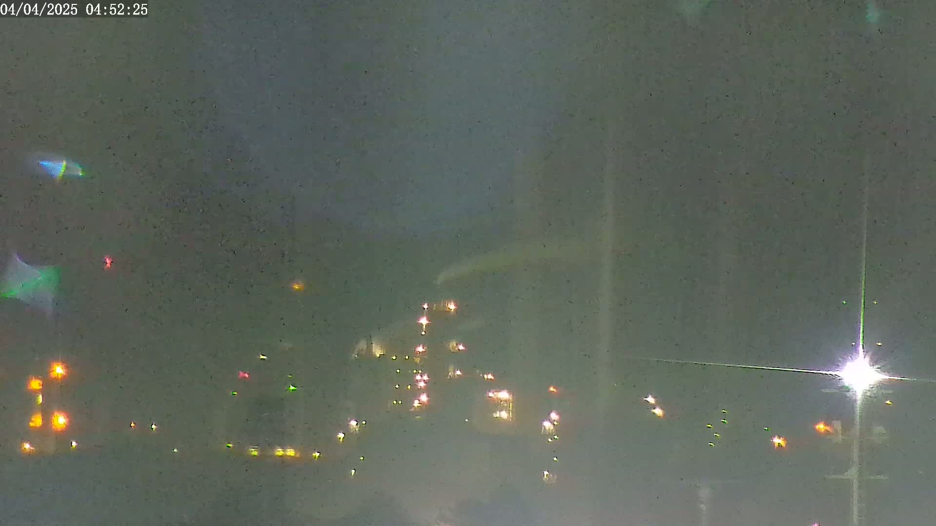 A nighttime, hazy view of a distant industrial complex illuminated by numerous lights, with a few additional lights visible in the foreground.