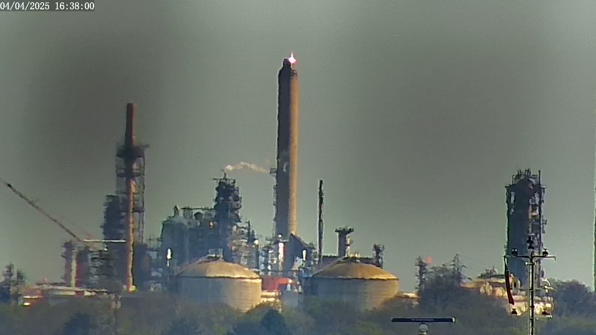 A large industrial refinery complex with tall smokestacks, some emitting smoke and one with a flame, is visible in hazy outdoor conditions.