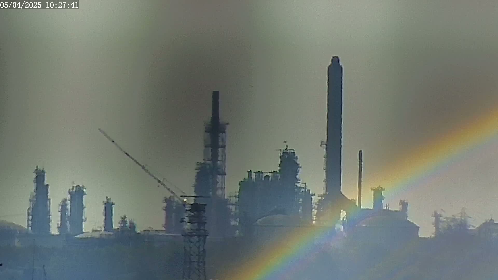 A partially visible rainbow arcs over a hazy industrial landscape of tall smokestacks and refinery structures.