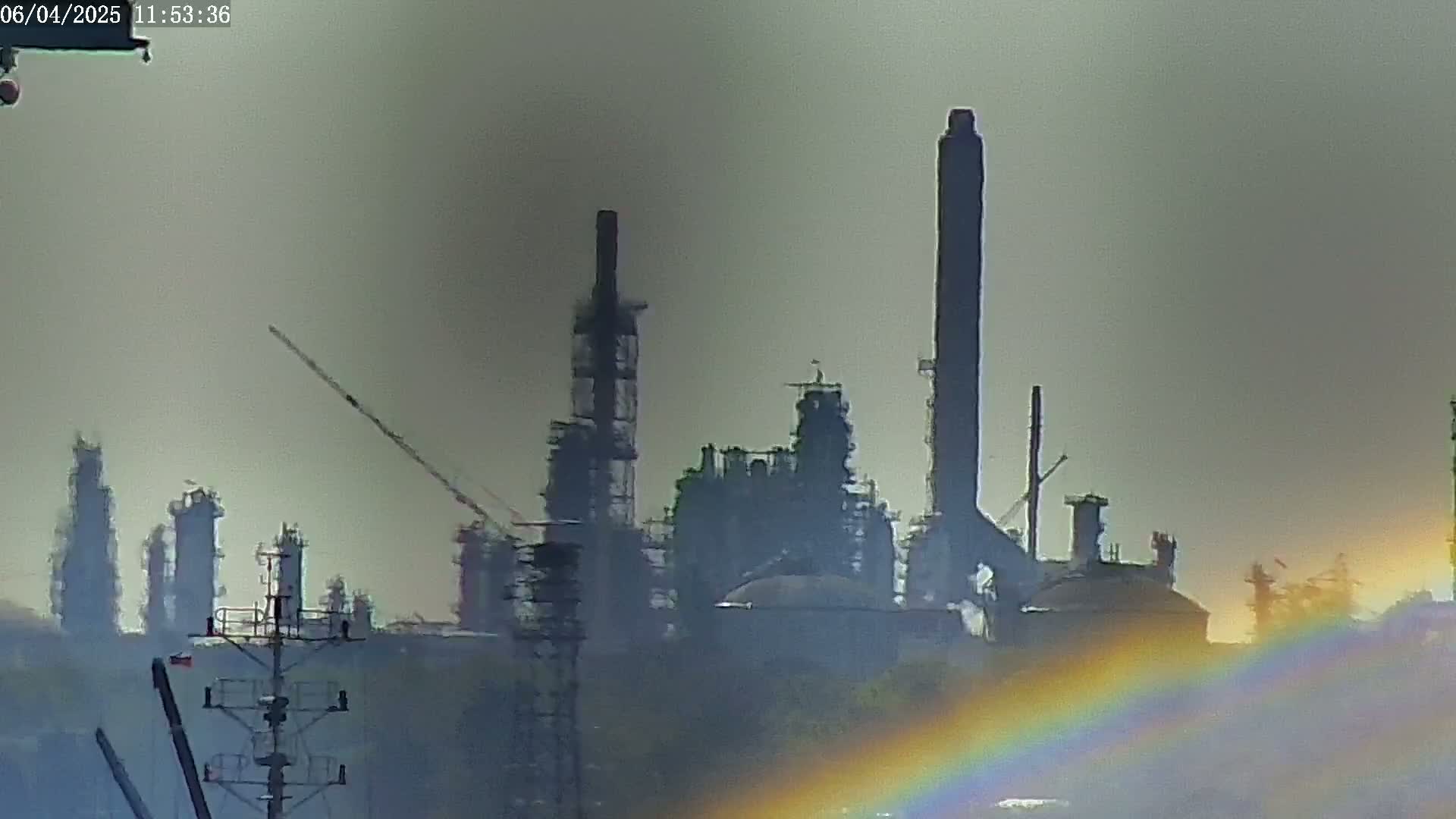 A hazy industrial complex with tall smokestacks is visible in the distance, partially obscured by a faint rainbow at the bottom.
