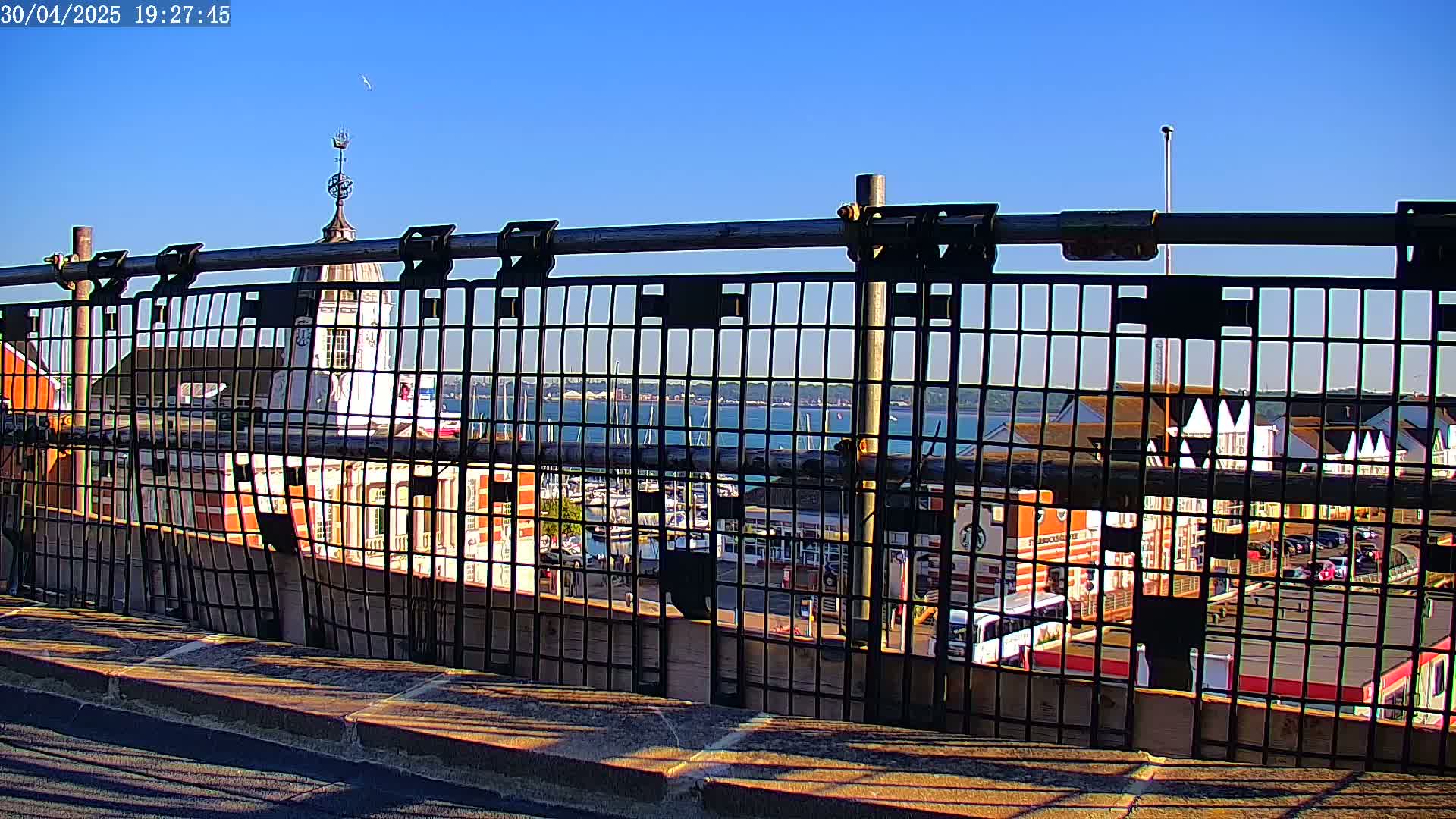 A metal fence obstructs the view of a harbor filled with boats, buildings, and a clear blue sky.