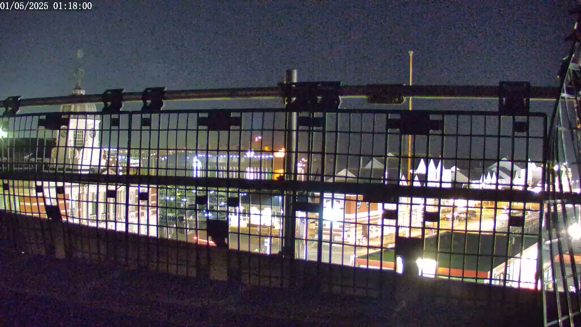 A nighttime view through a metal railing shows a brightly lit waterfront area with buildings and a pier.