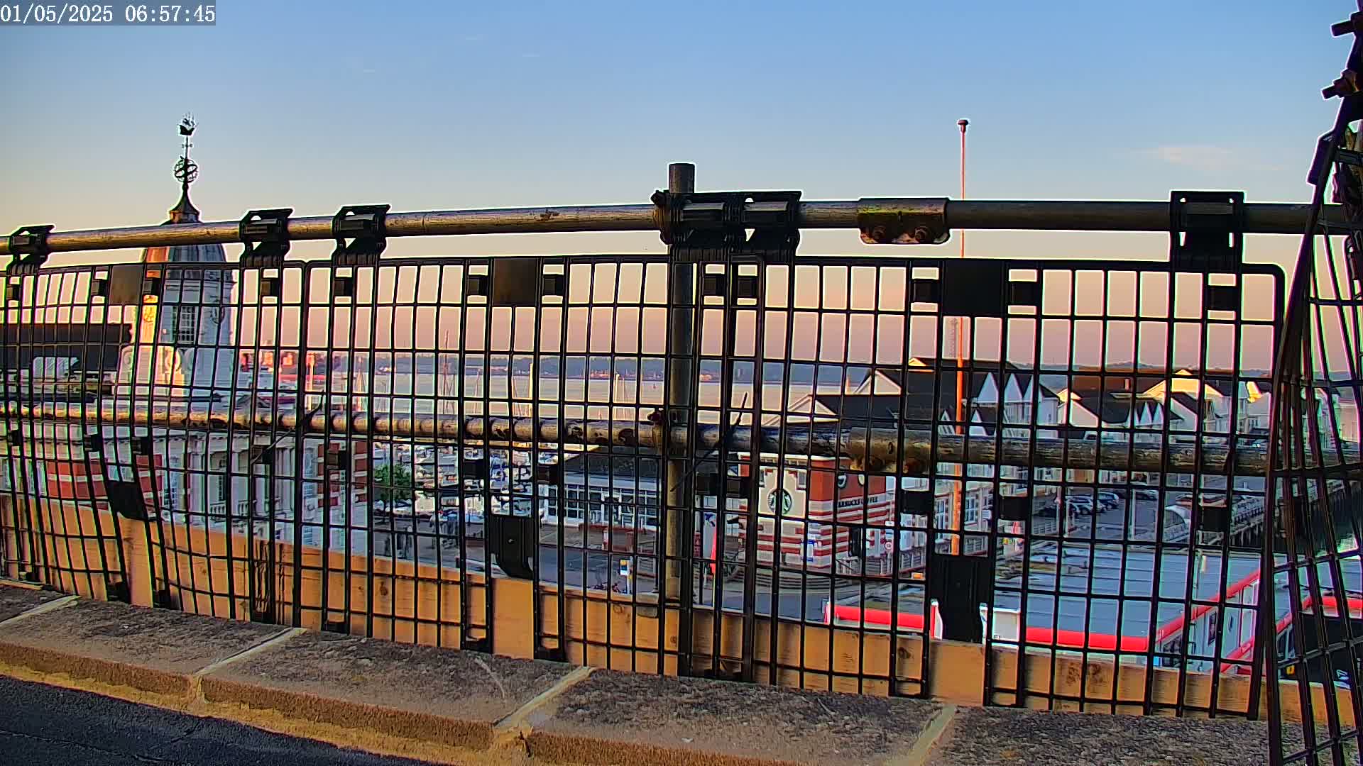 A metal railing overlooks a harbor at sunset, showing a town with buildings and boats.