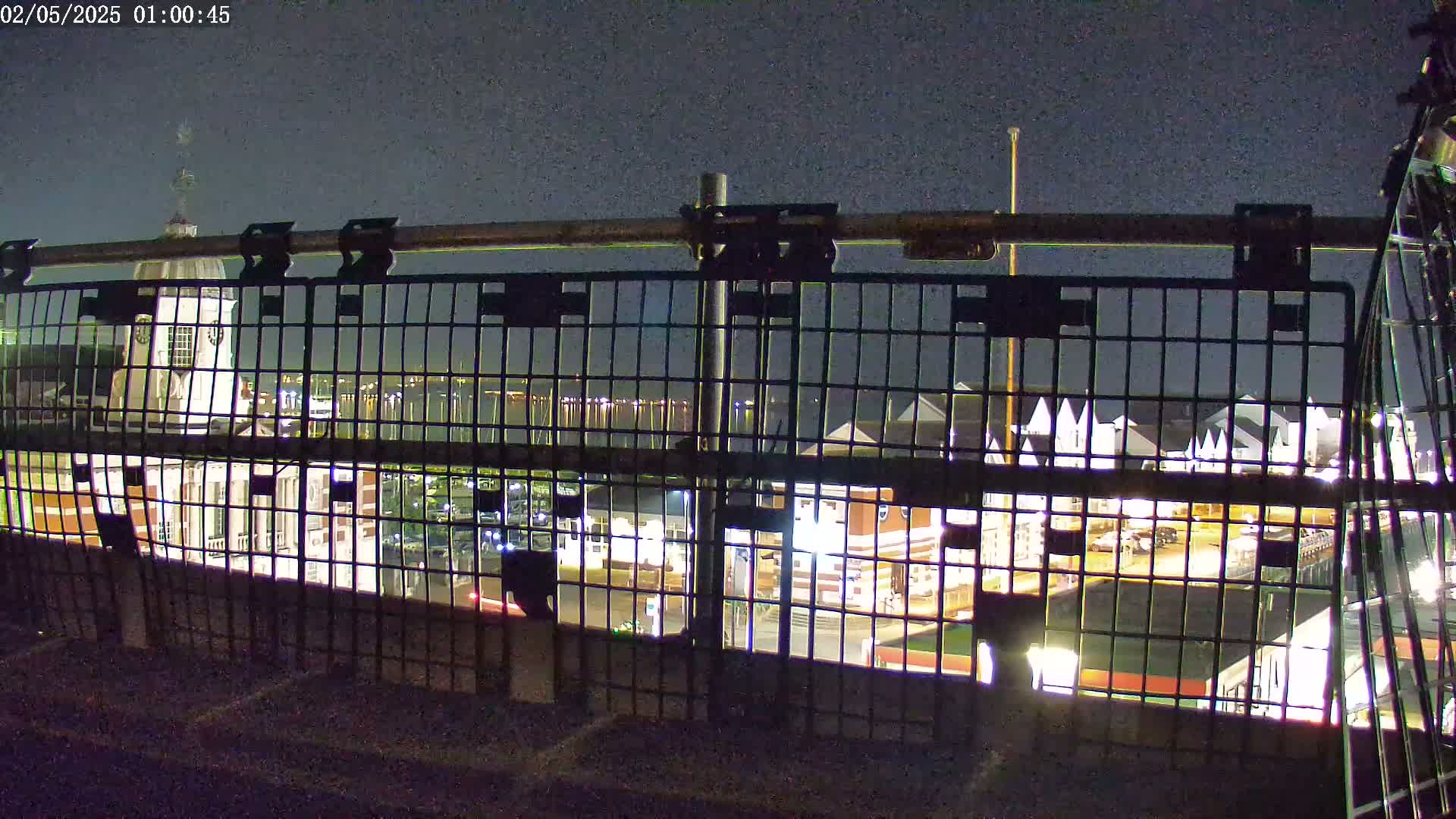 A nighttime view through a metal railing shows a waterfront area with buildings and boats, under a dark sky.