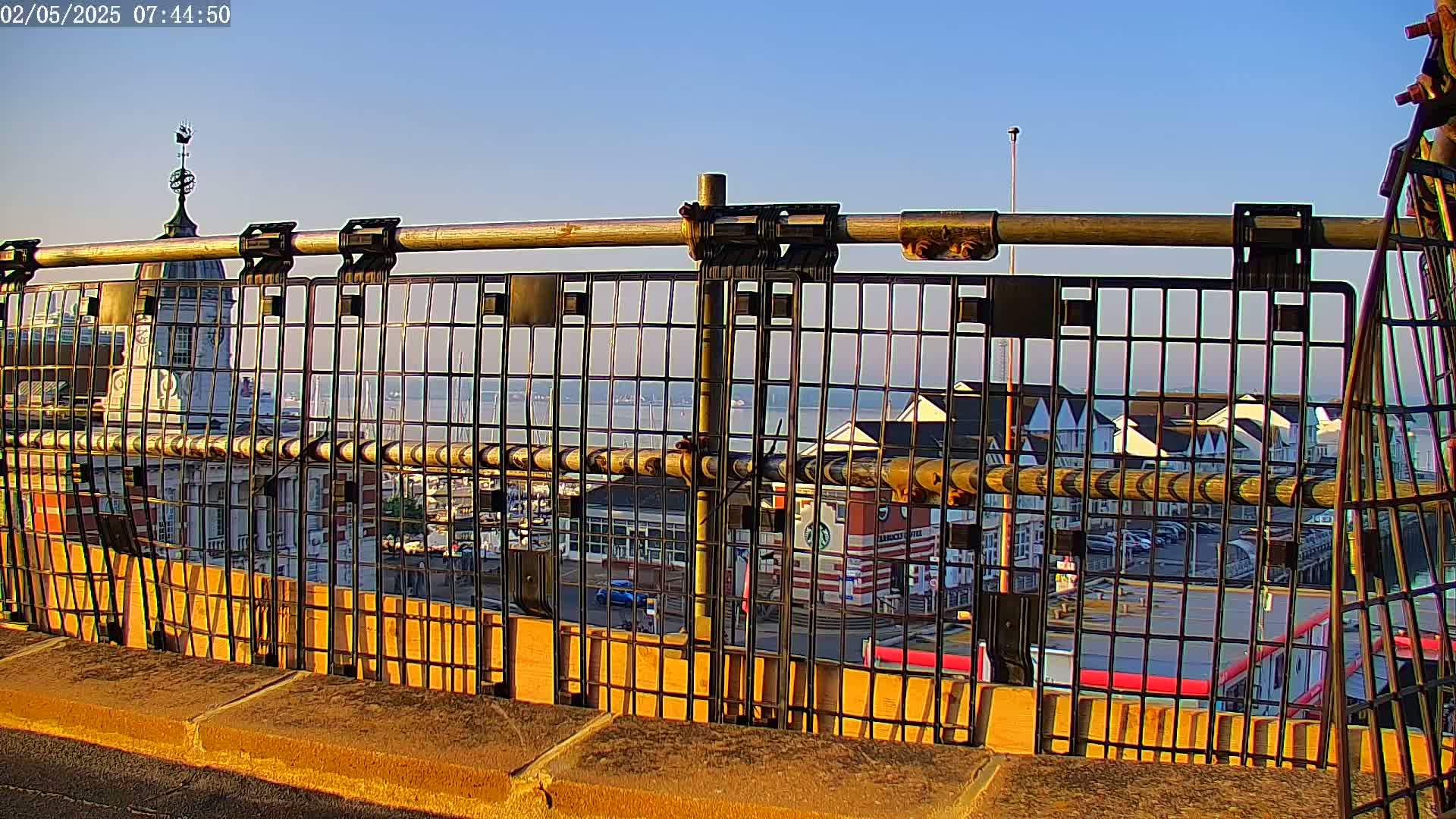 A metal railing with a view of a harbor, buildings, and calm water under a clear blue sky.