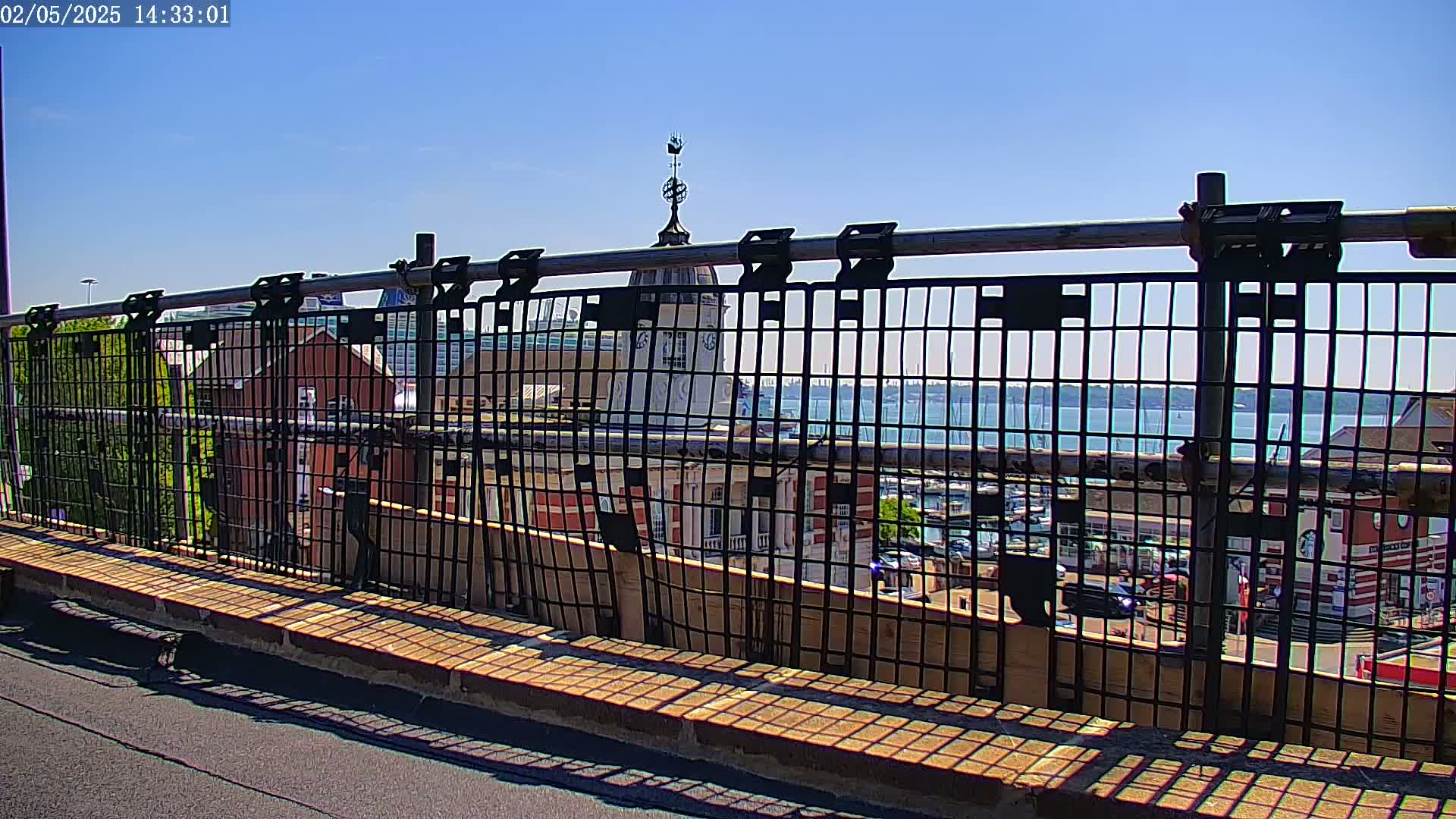 A metal railing on a sunny day provides a view of a body of water with boats, buildings, and trees.