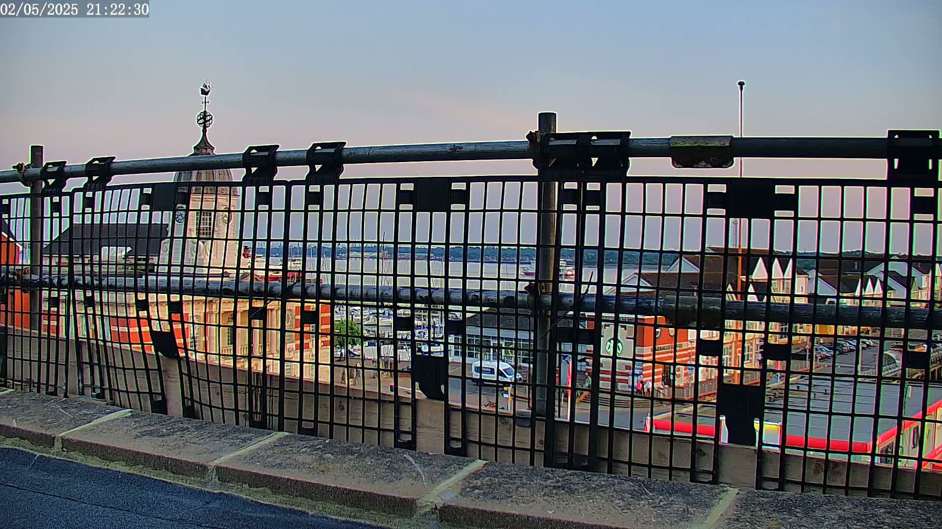 A metal fence provides a view of a harbor with boats, buildings, and a calm evening sky.