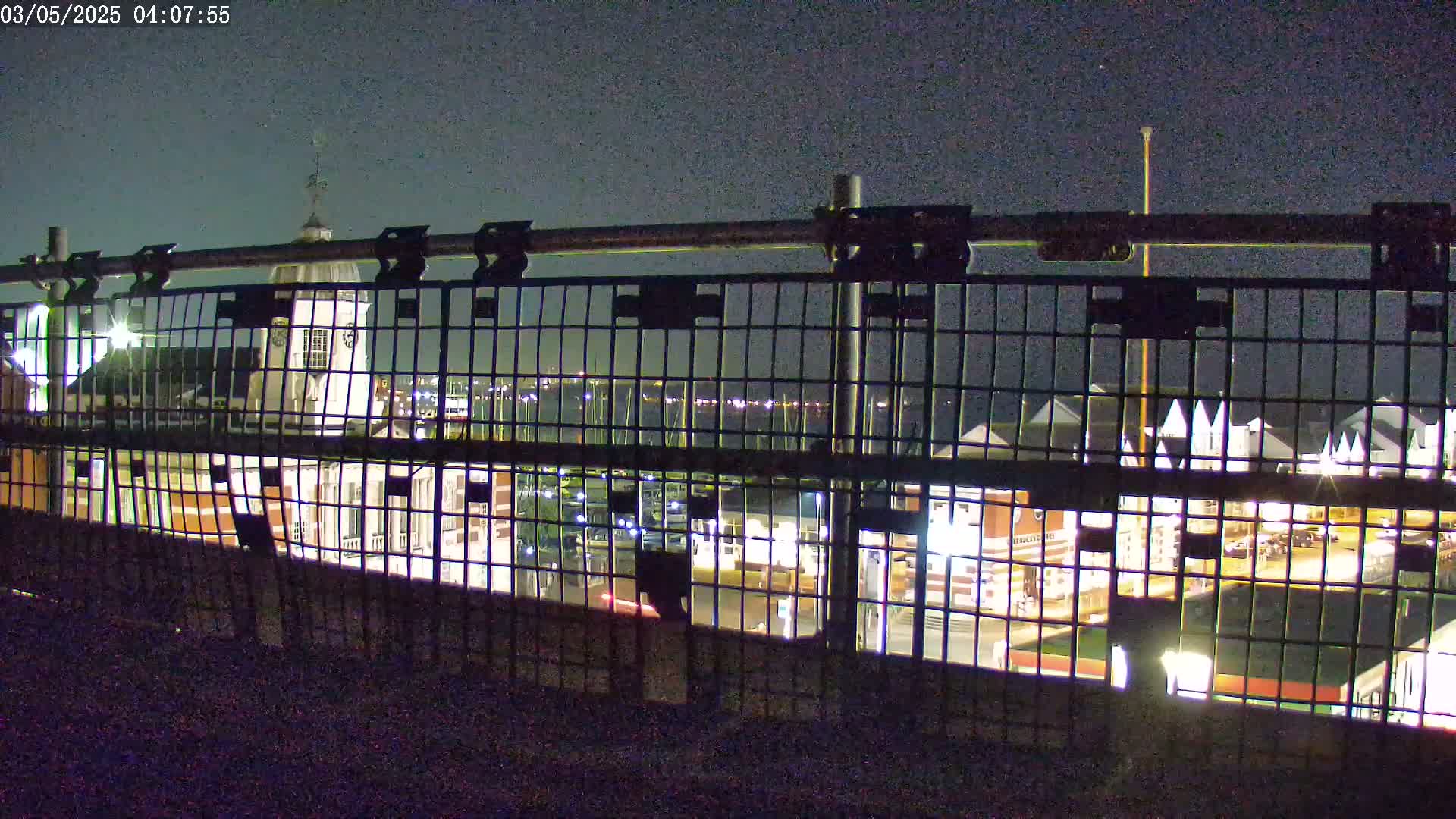 A nighttime view of a harbor and buildings, seen through a metal fence under a dark sky.