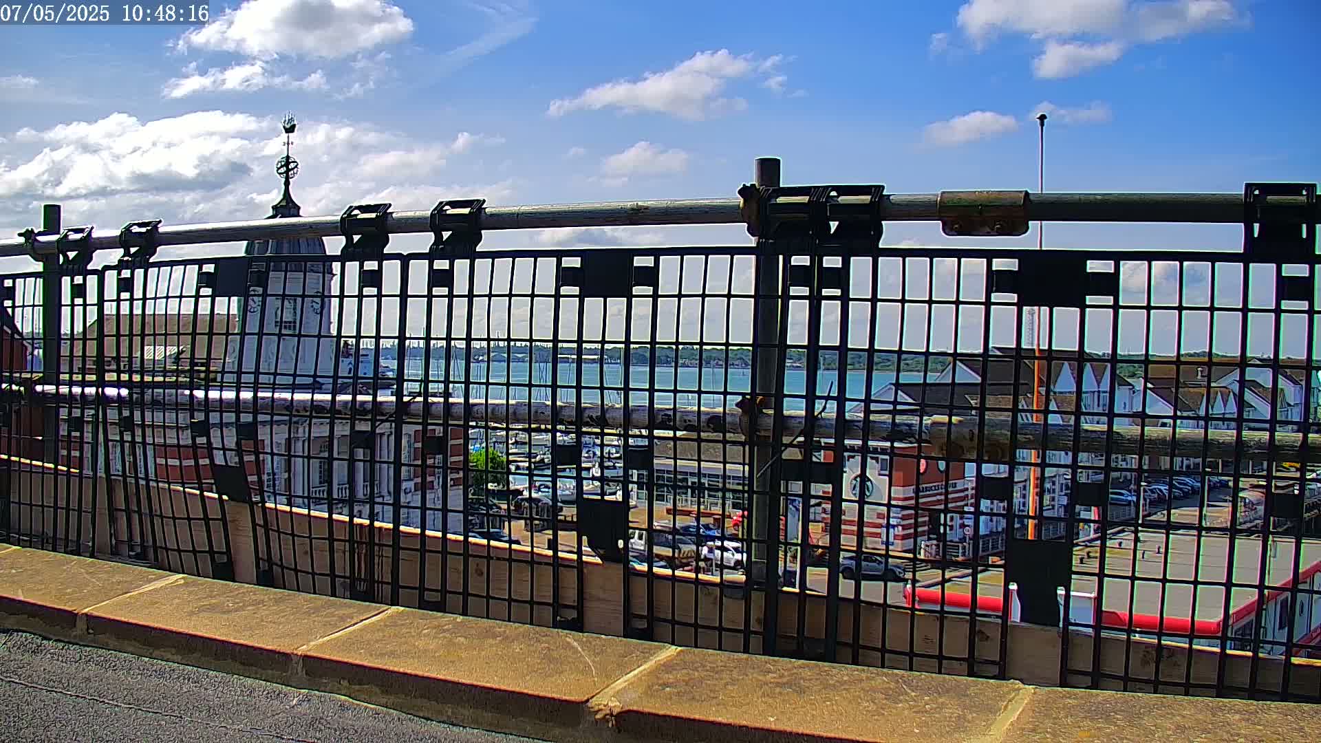 A metal fence overlooks a harbor with boats, buildings, and a partly cloudy sky.