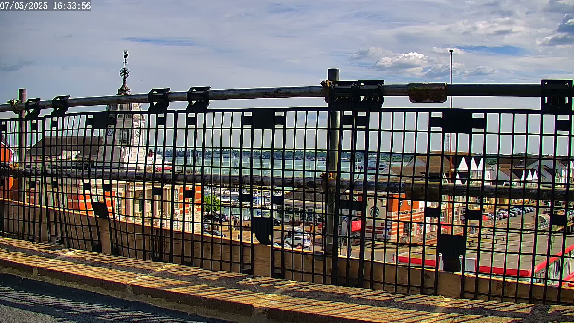 A partly cloudy sky is visible over a waterfront town seen through a metal fence, with a clock tower prominent in the background.