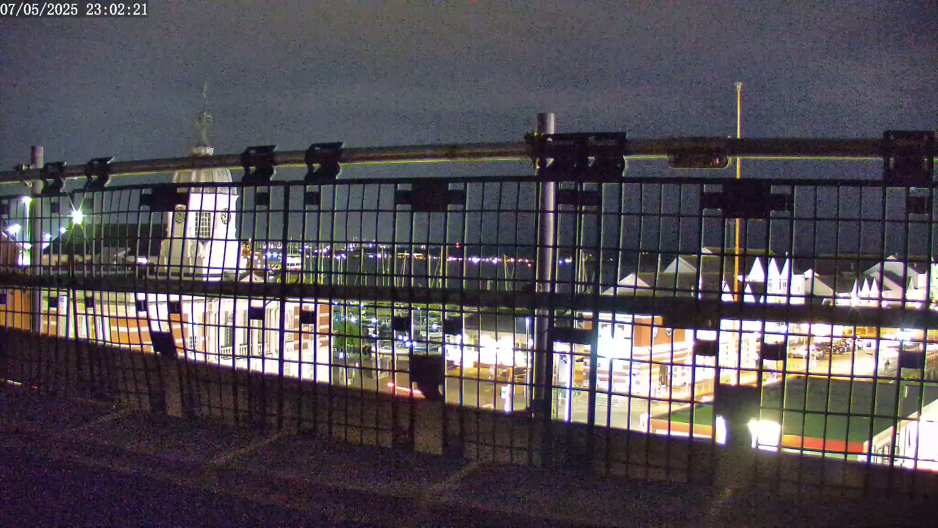 A nighttime view of a marina, seen through a metal fence, under a dark sky.