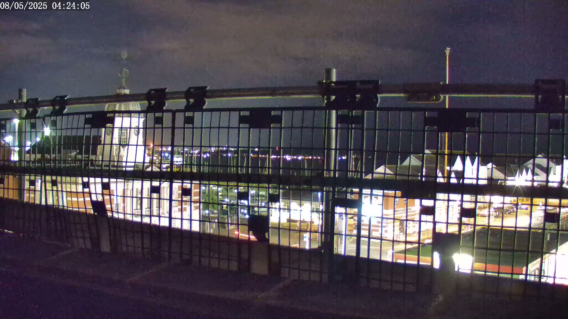 A nighttime view through a metal fence shows a building with a cupola and other illuminated structures, all under a partly cloudy sky.