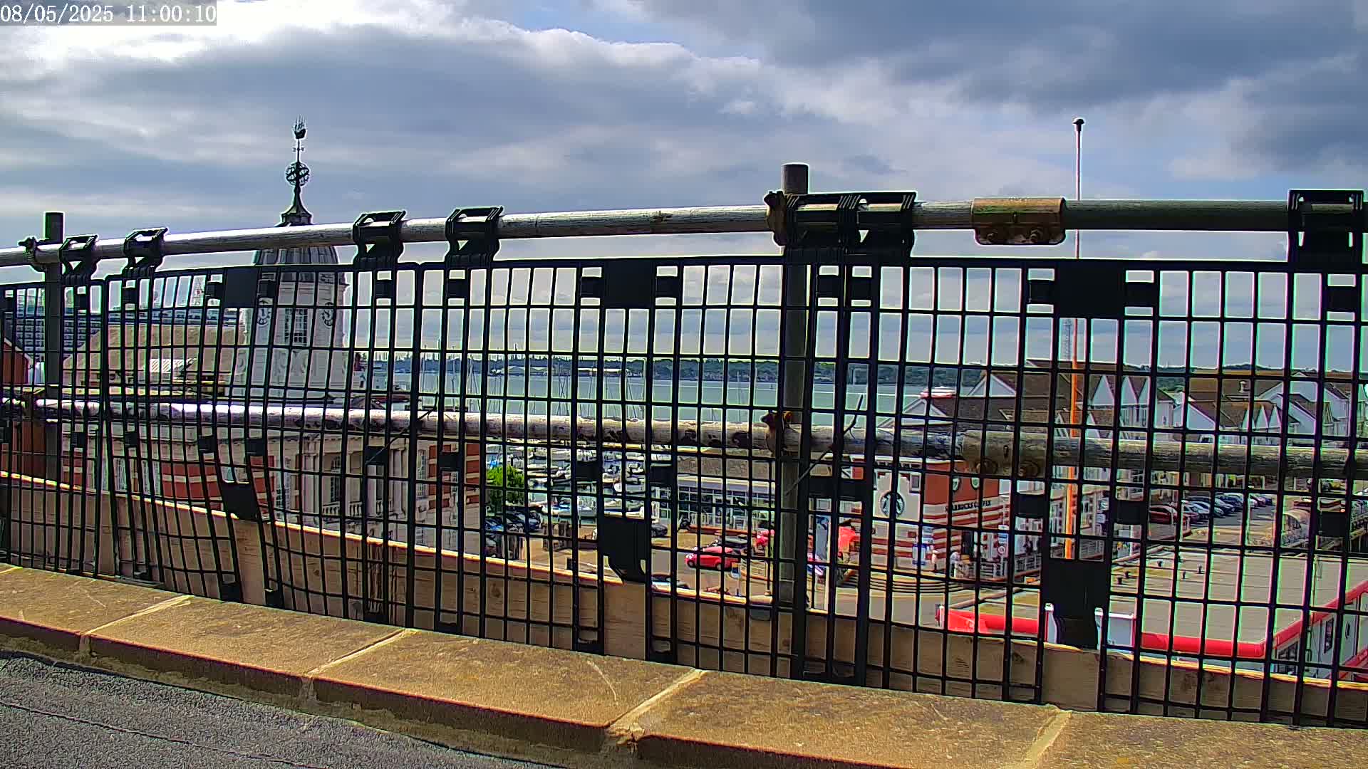 A partially cloudy sky is visible over a body of water filled with boats, seen through a black metal fence atop a stone wall.