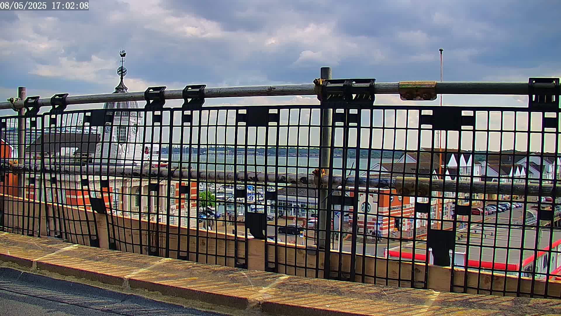A partially constructed metal railing on a rooftop overlooks a harbor with boats, buildings, and a partly cloudy sky.