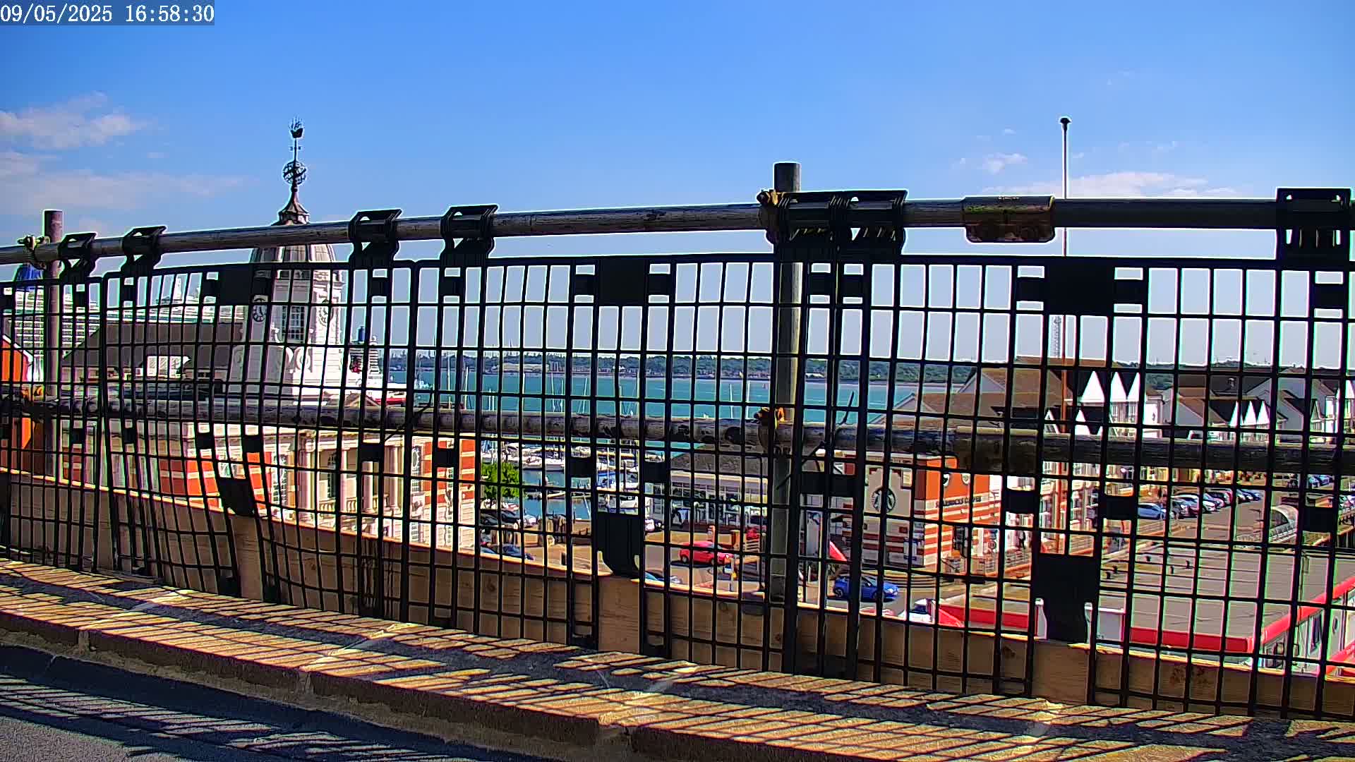 A metal fence obstructs the view of a harbor with boats and buildings on a sunny day.