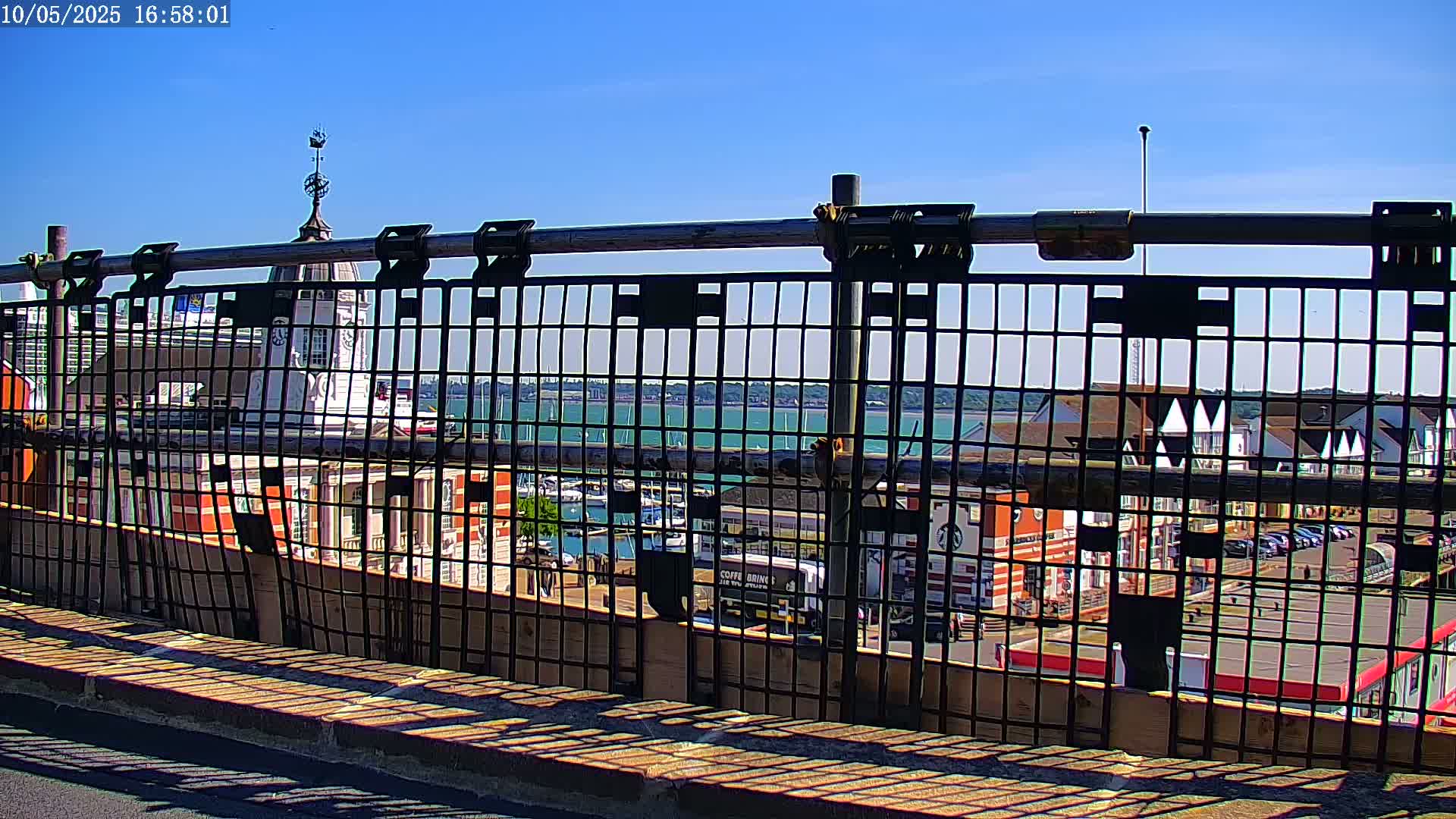 A metal fence casts shadows on a paved surface, offering a view of a harbor filled with boats under a clear blue sky.