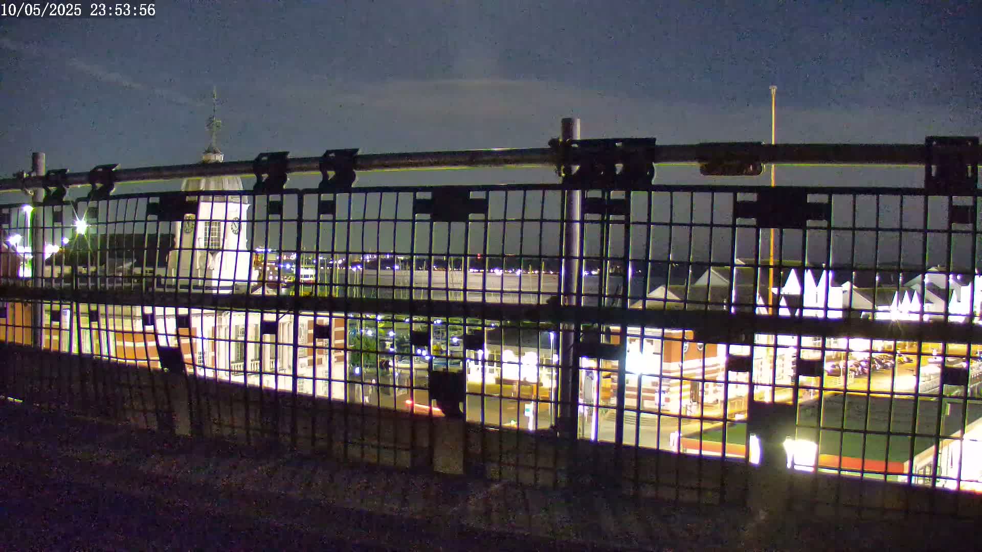 A nighttime view through a metal fence shows a waterfront area with illuminated buildings and a body of water beyond.
