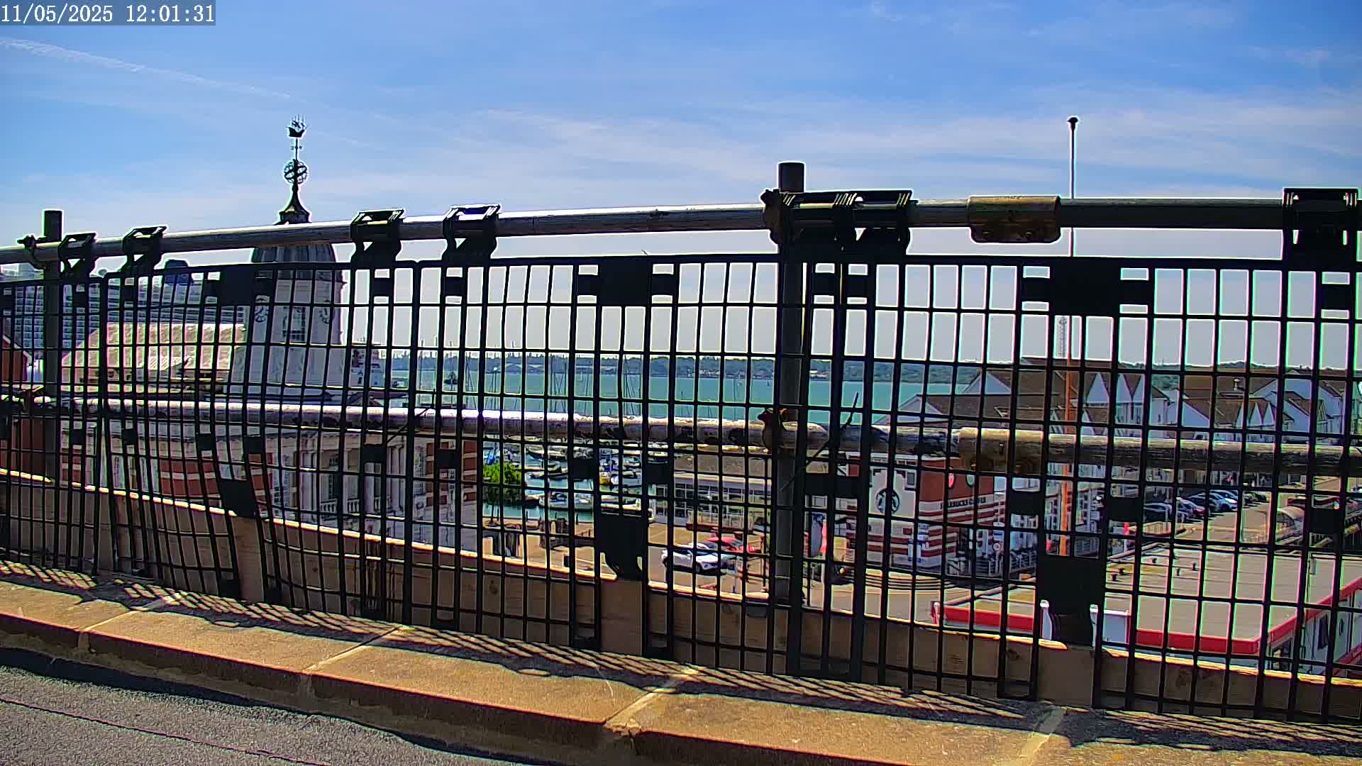 A black metal fence provides a view of a harbor filled with boats on a sunny day.