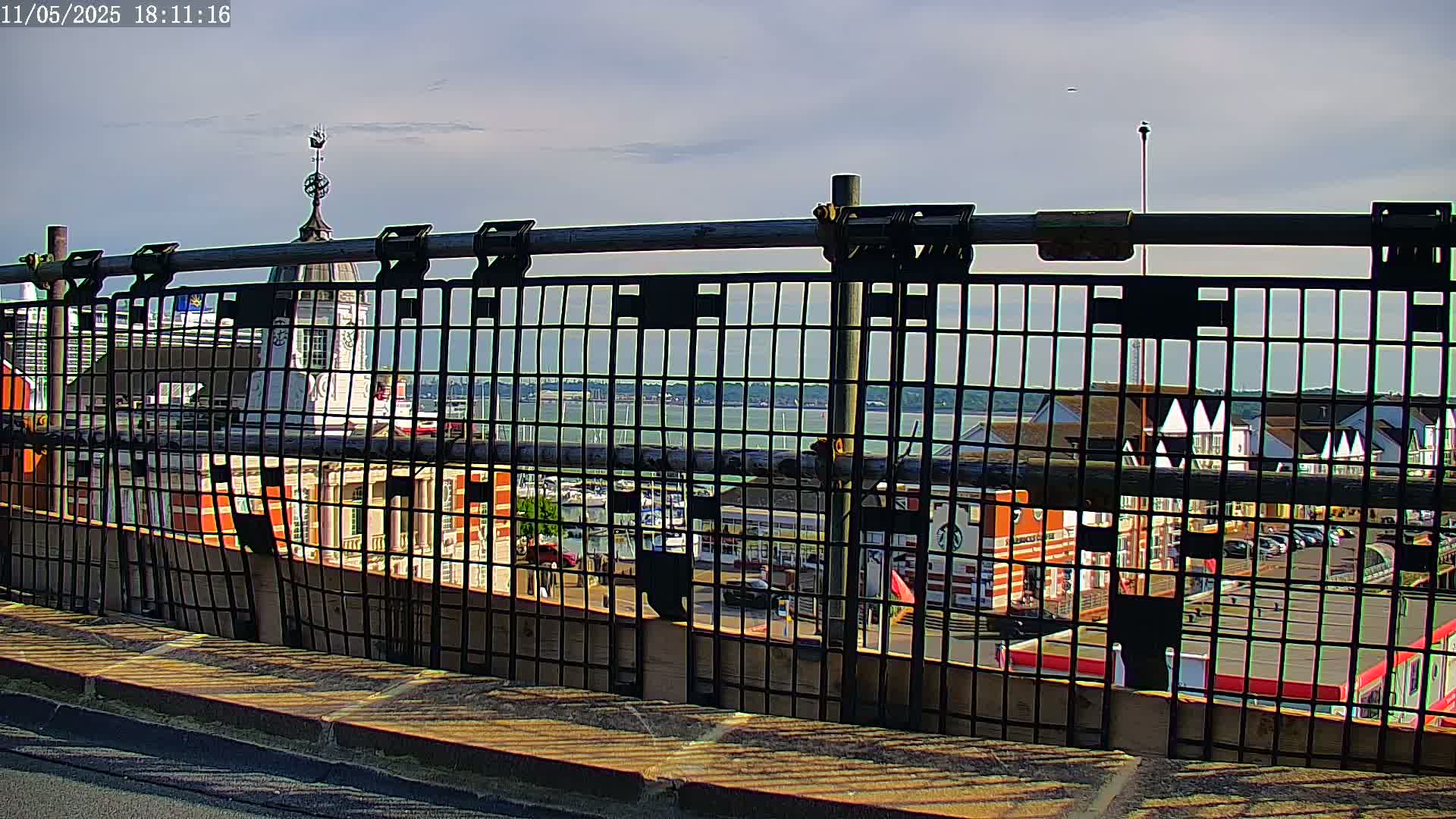 A metal fence with a view of a waterfront area with buildings, boats, and a partly cloudy sky.