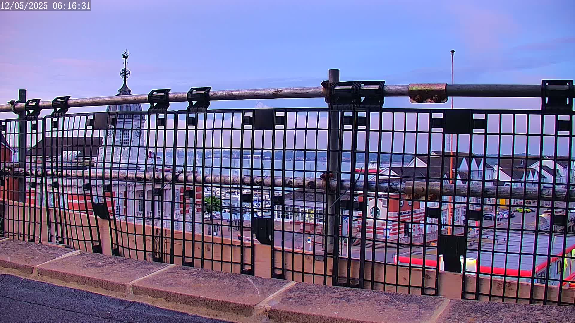 A metal fence frames a view of a harbor town under a mostly clear, light-purple sky.