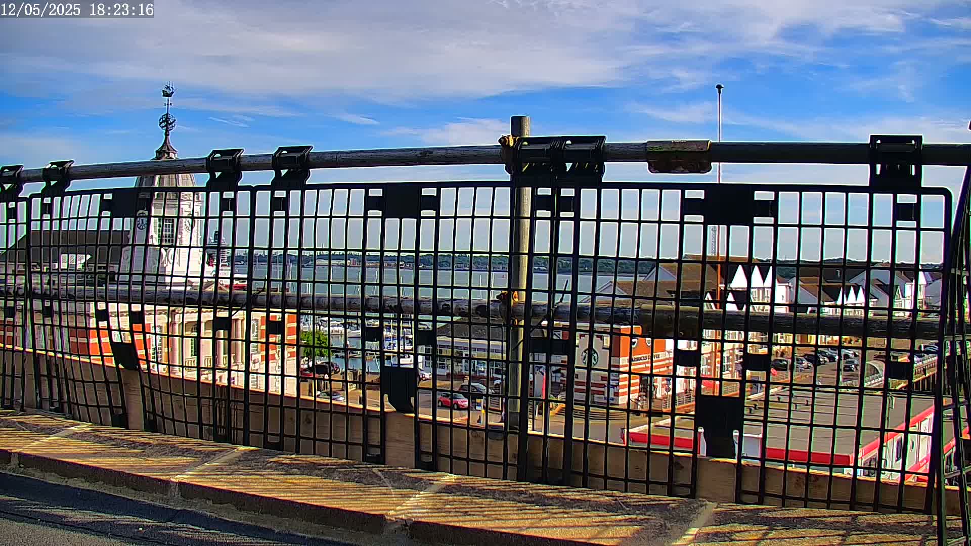 A metal fence provides a view of a harbor, buildings, and a body of water under a mostly sunny sky.