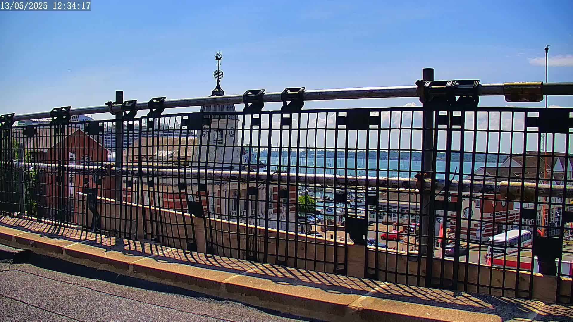 A metal fence on a sunny day provides a view of a harbor with boats, buildings, and vehicles.