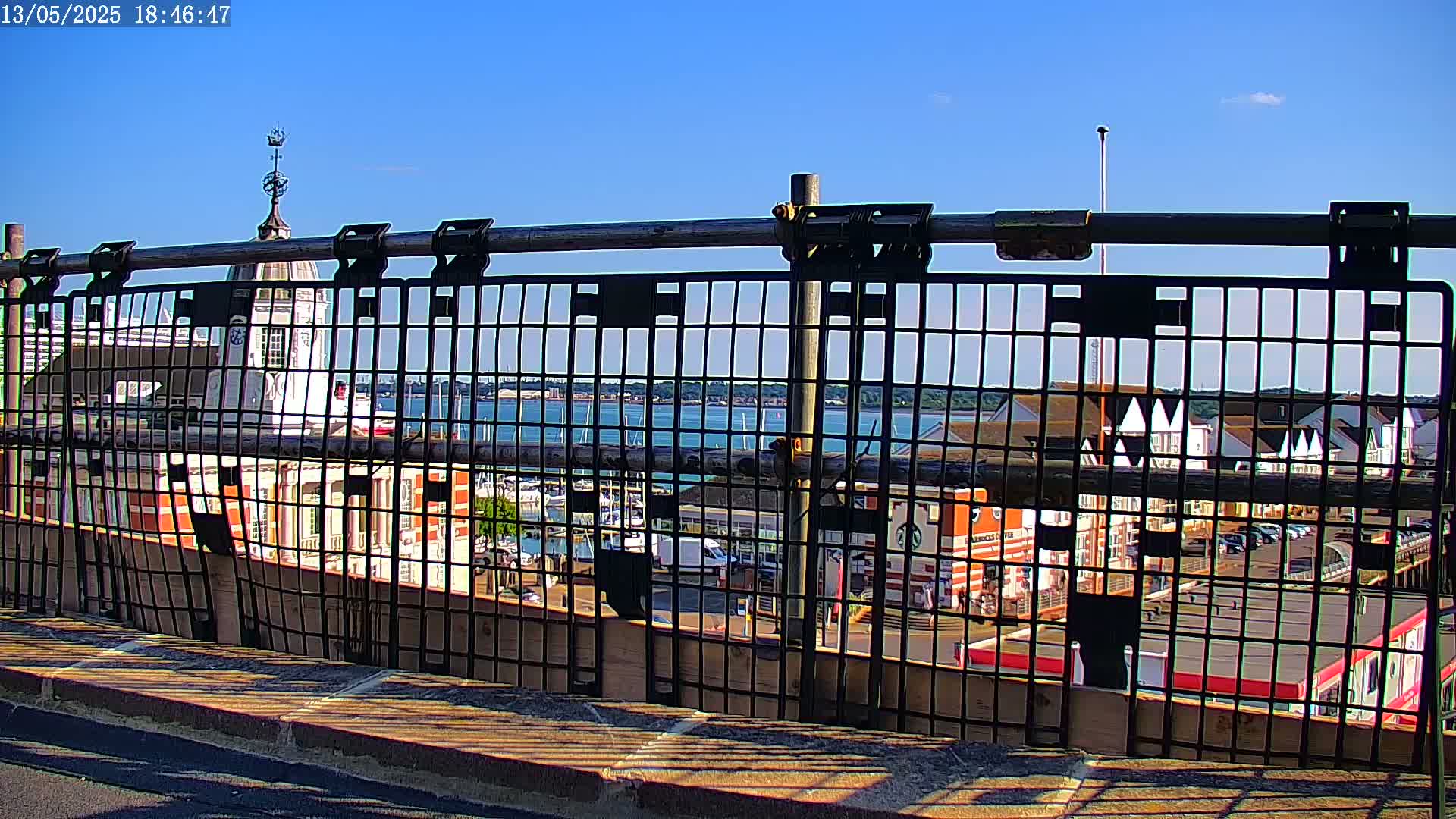 A metal grid fence partially obscures a view of a waterfront scene on a sunny day, featuring buildings, boats, and a body of water.