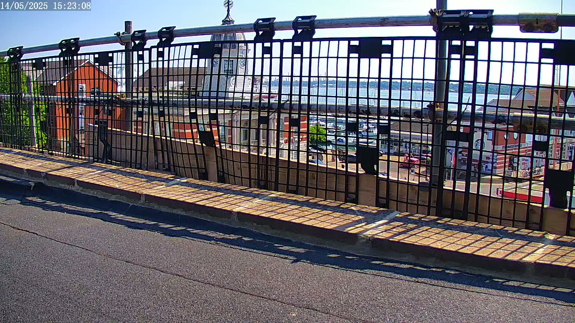A black metal fence on a sunny day overlooks a harbor with boats, buildings, and a roadway below.