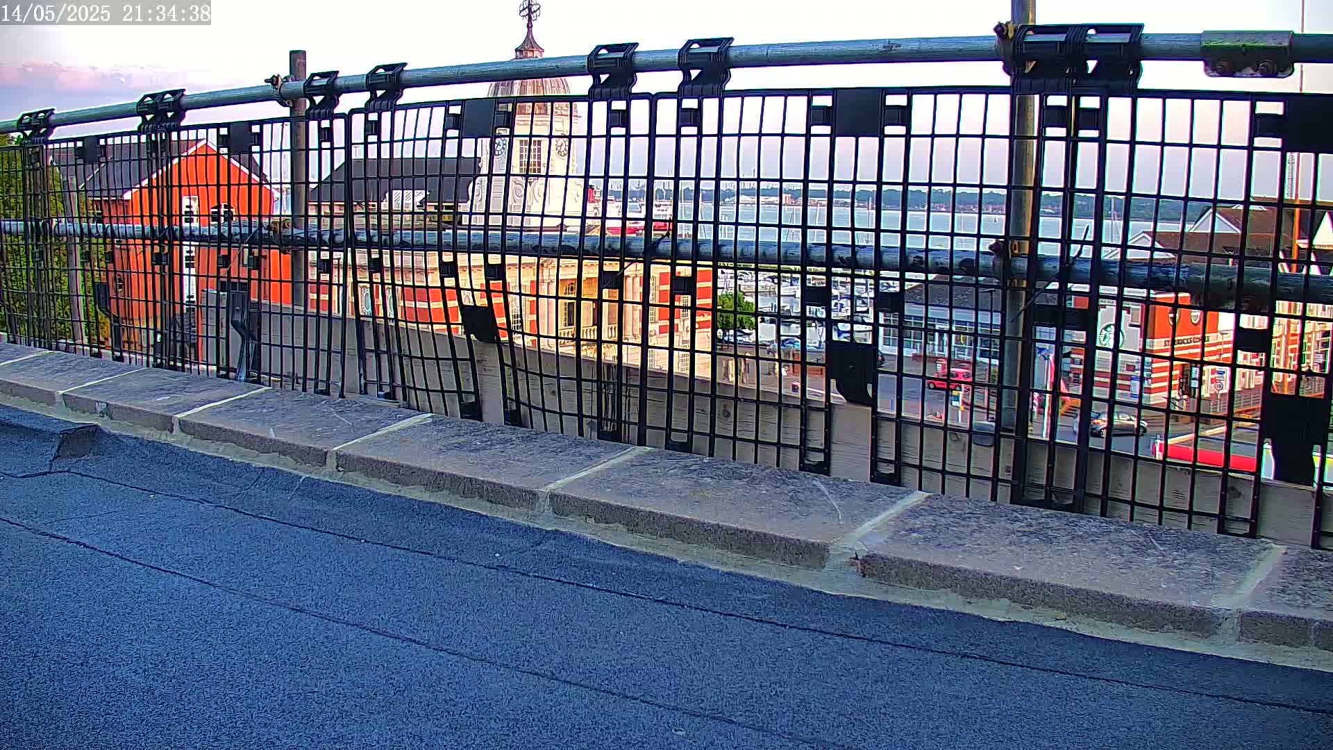 A black metal fence on a rooftop overlooks a harbor scene with a red brick building and other structures at dusk.