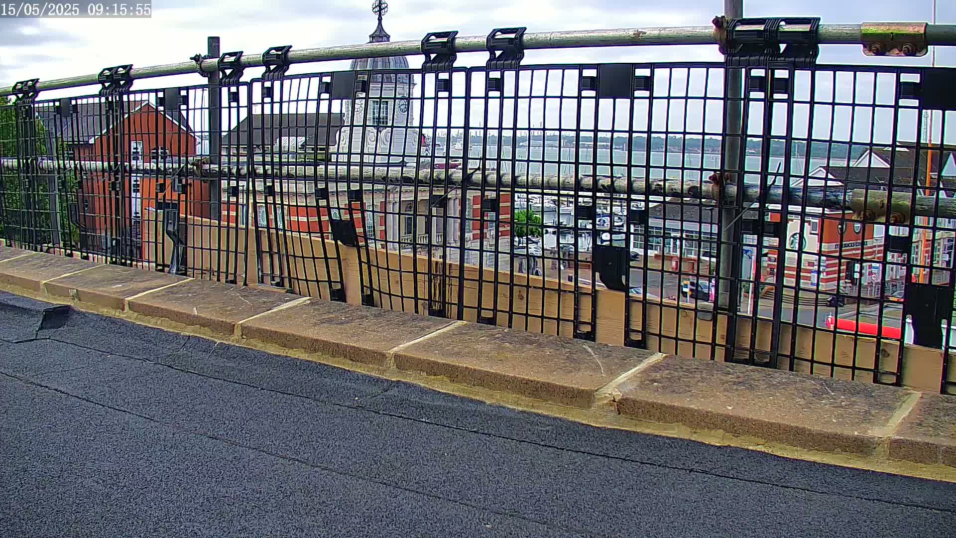 A black metal fence with a view of a town, a body of water, and a cloudy sky.
